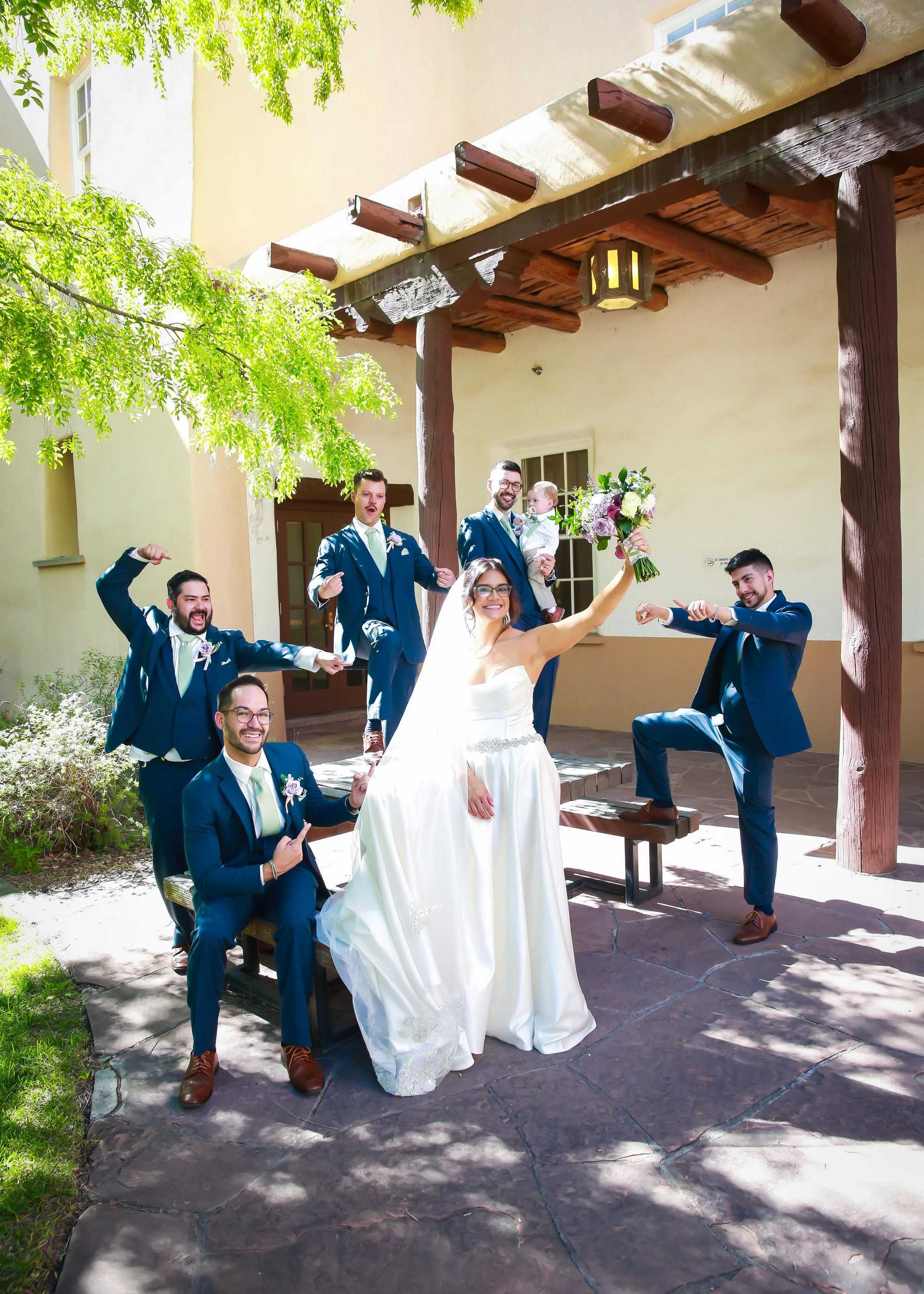 Group of people celebrating at a wedding outdoors, with the bride holding a bouquet and the groom and friends making playful poses.