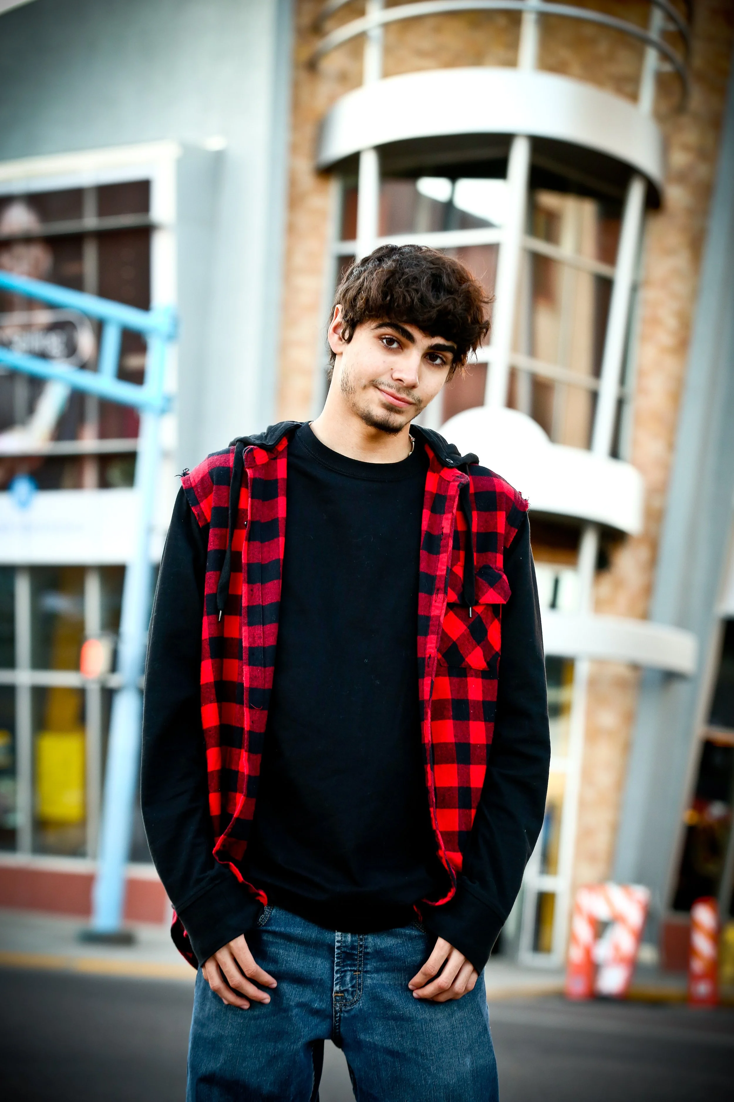 A young man standing on a city street in front of a building with large industrial-style windows and a spiral staircase. He is wearing a black t-shirt, a red and black plaid vest, and blue jeans, with hands in his pockets.