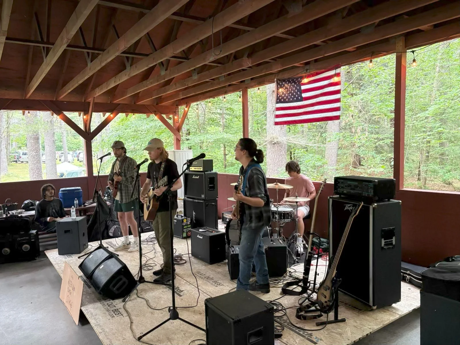 Maintenance musicians performing on a covered outdoor stage surrounded by trees, with American flag hanging in the background.