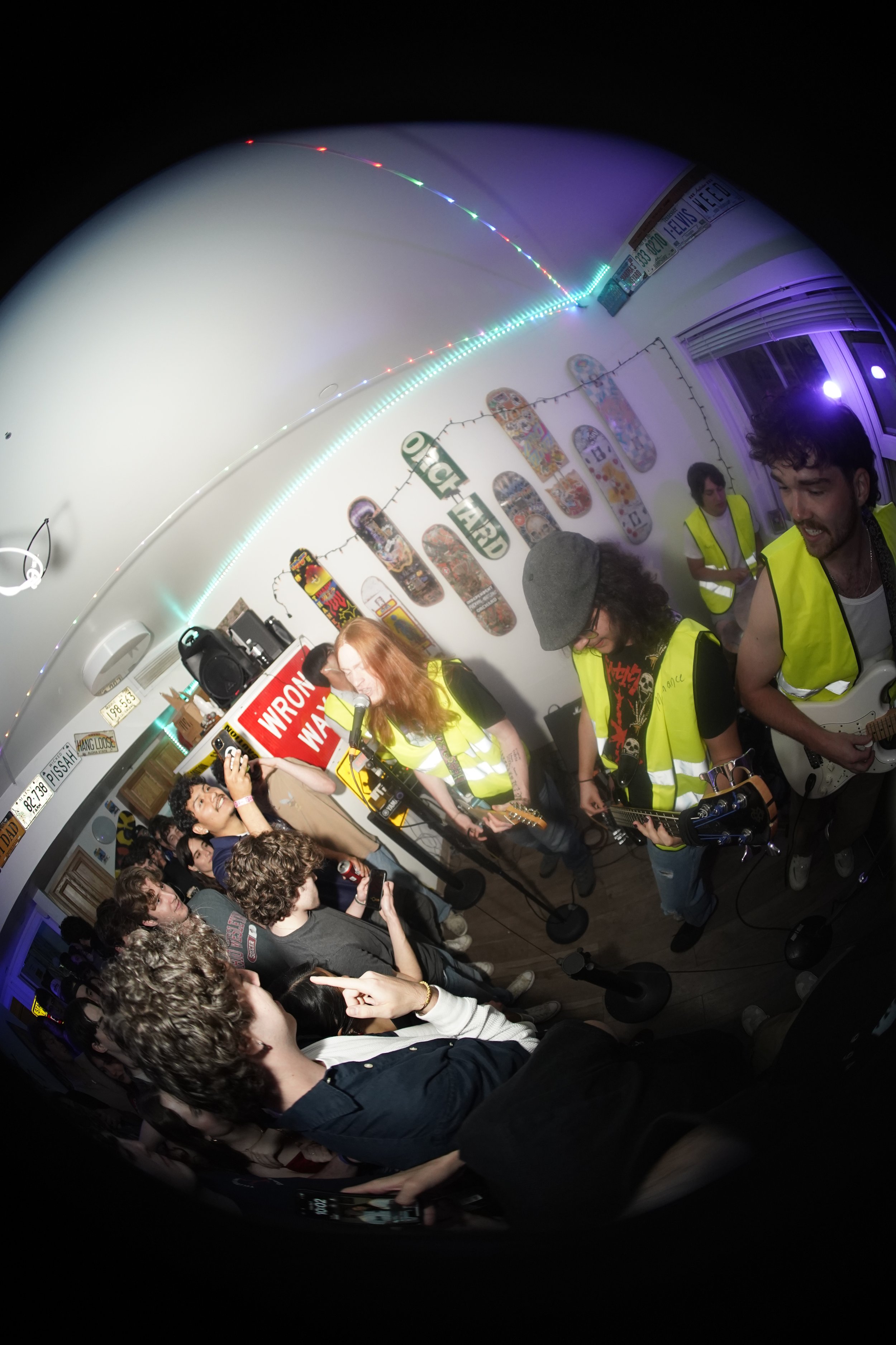 Maintenance performance at a small indoor venue with a crowd of people watching. The band members are wearing yellow safety vests, with one playing guitar and another singing into a microphone.