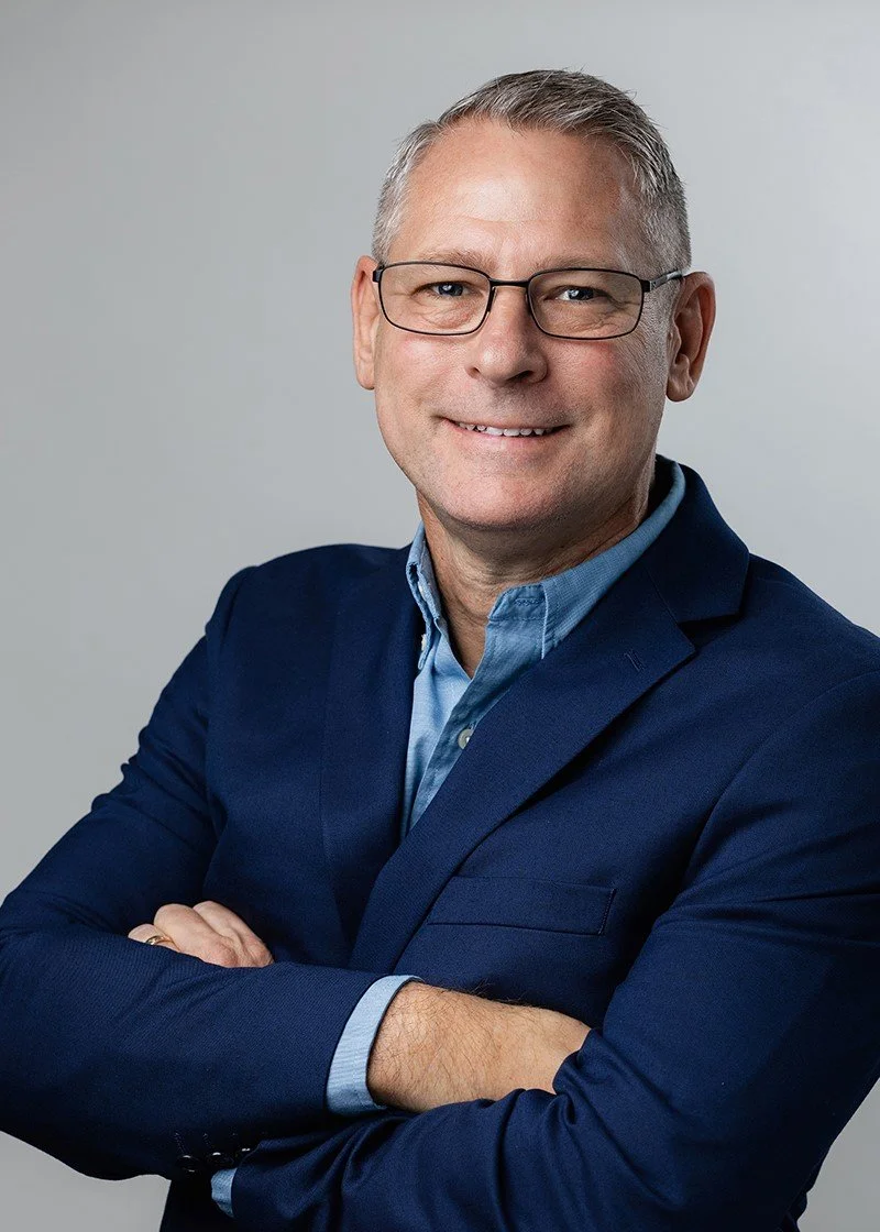 Portrait of a middle-aged man with glasses wearing a navy blue blazer and light blue shirt, arms crossed, smiling against a light gray background.
