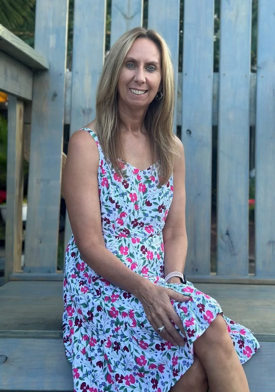 A woman with long blonde hair, wearing a sleeveless white dress with pink, red, purple, and green floral pattern, sitting on wooden steps in front of a wooden fence, smiling at the camera.