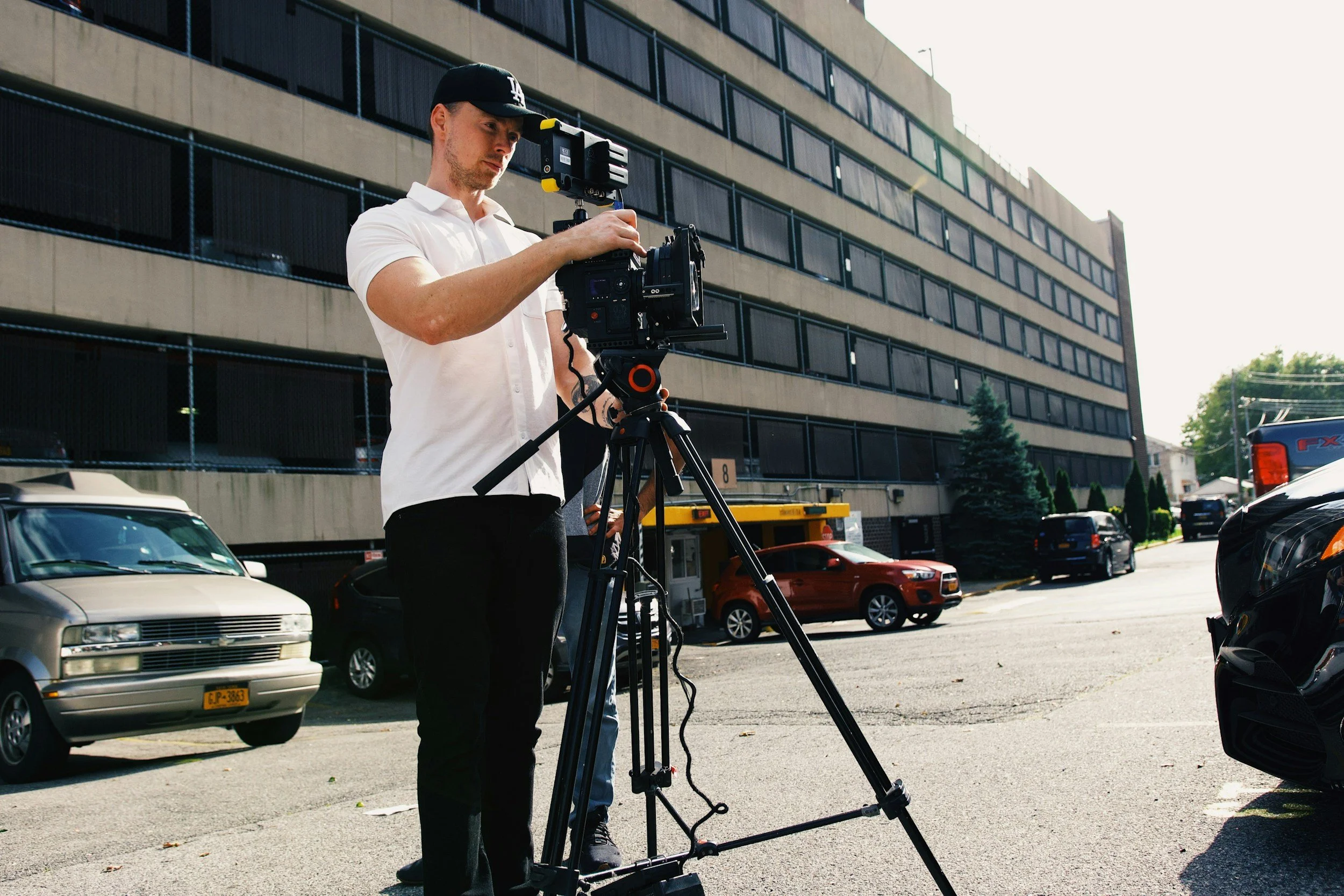 A man wearing a black cap and white shirt operating a professional camera on a tripod in a parking lot with several cars and a multi-story building in the background.