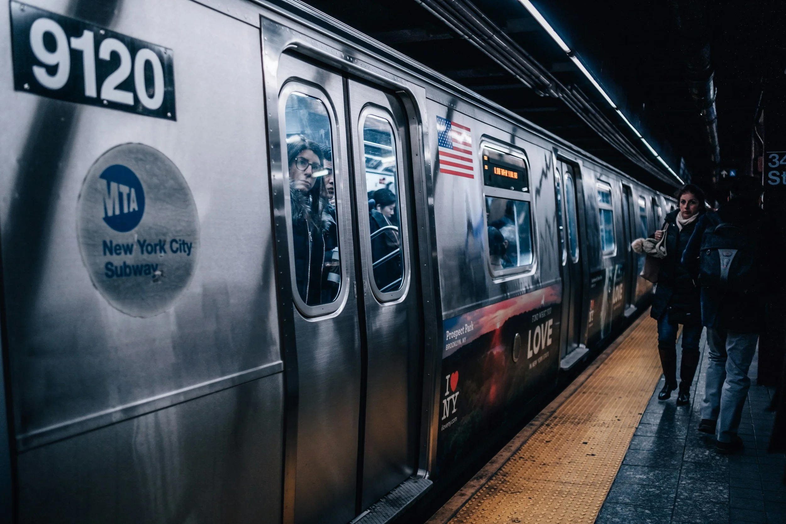 People waiting on the platform for a New York City subway train, with reflections in the train's windows, an American flag sticker, and an electronic sign displaying train information.