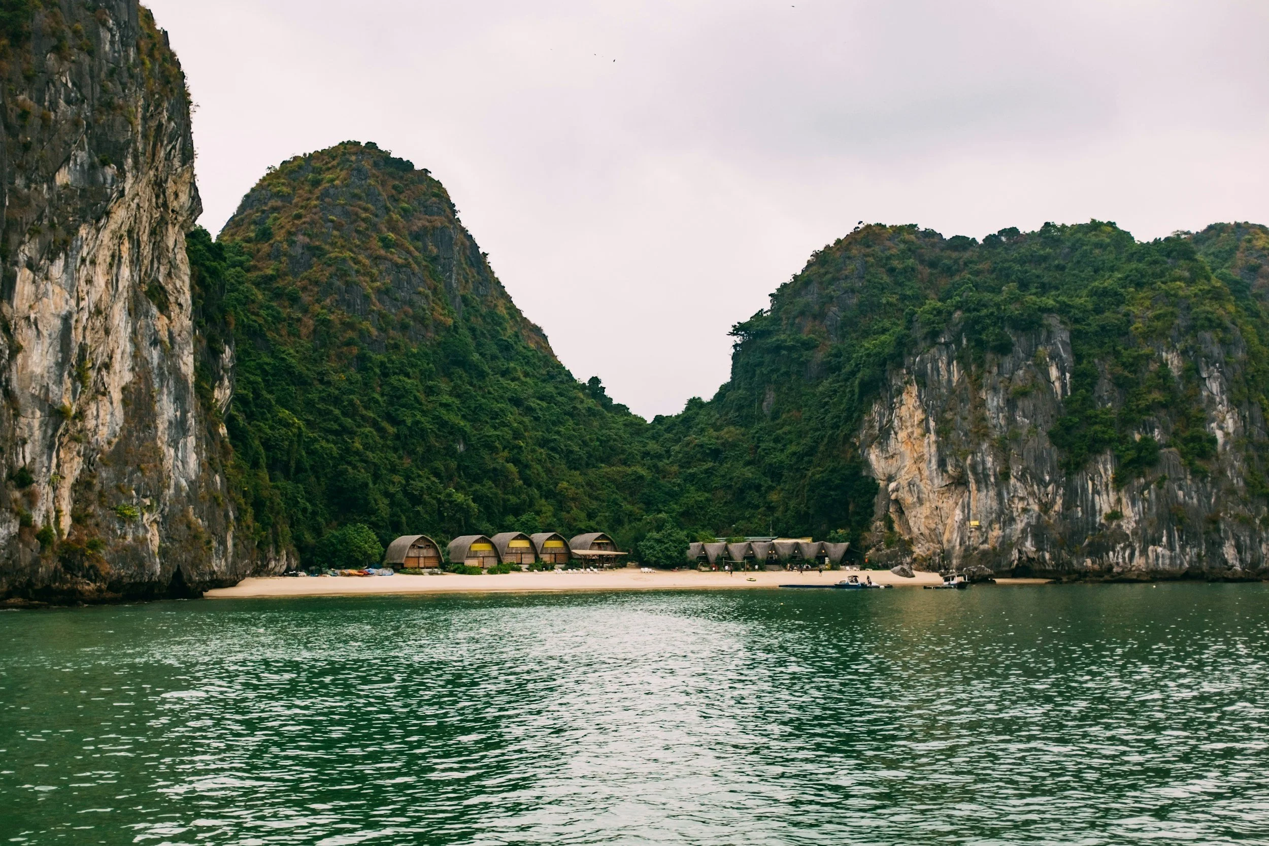 Tropical beach with tall cliffs and lush green vegetation, small huts on the sandy shore, and calm water in the foreground, under an overcast sky.