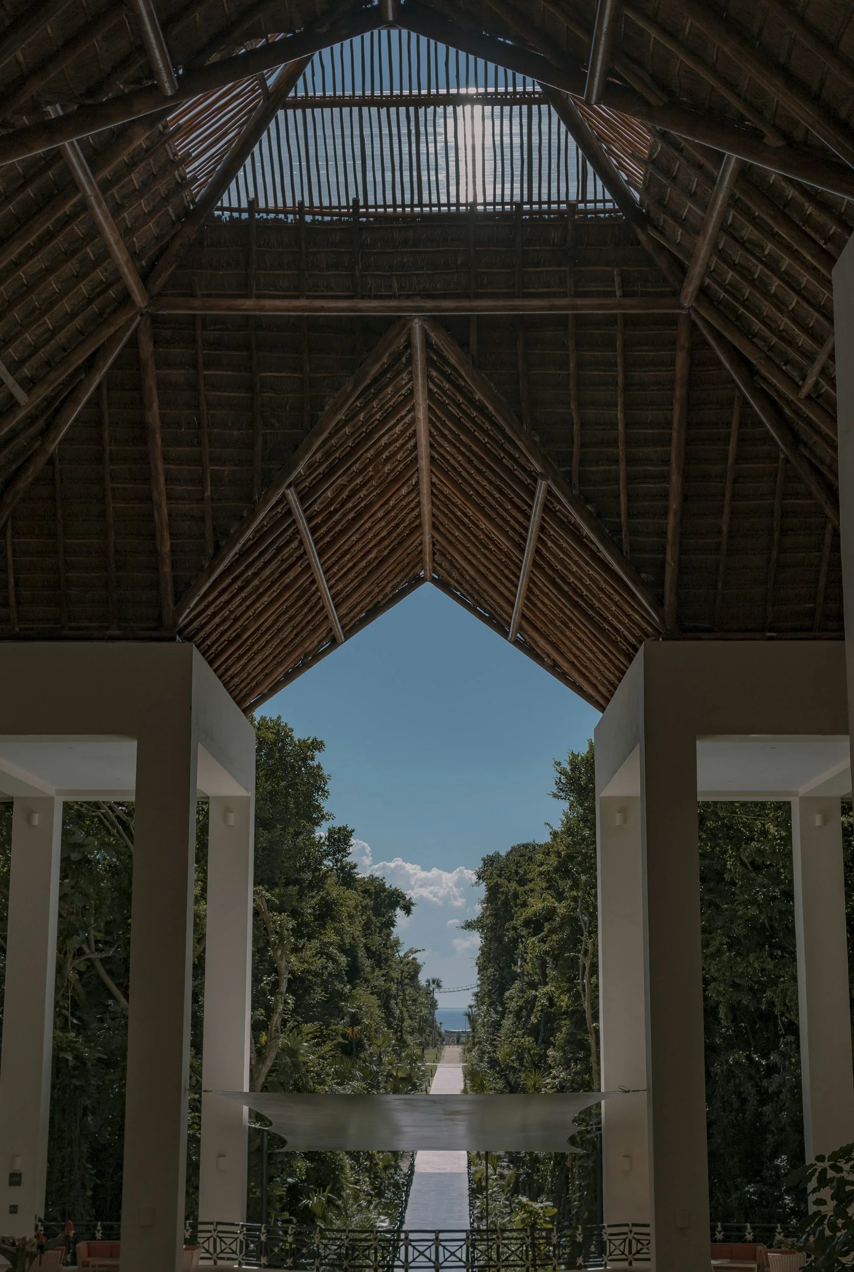 Looking up at the high ceiling of a building with a bamboo or thatch roof. Outside, a pathway lined with trees and greenery extends toward the horizon with water and sky visible in the distance.