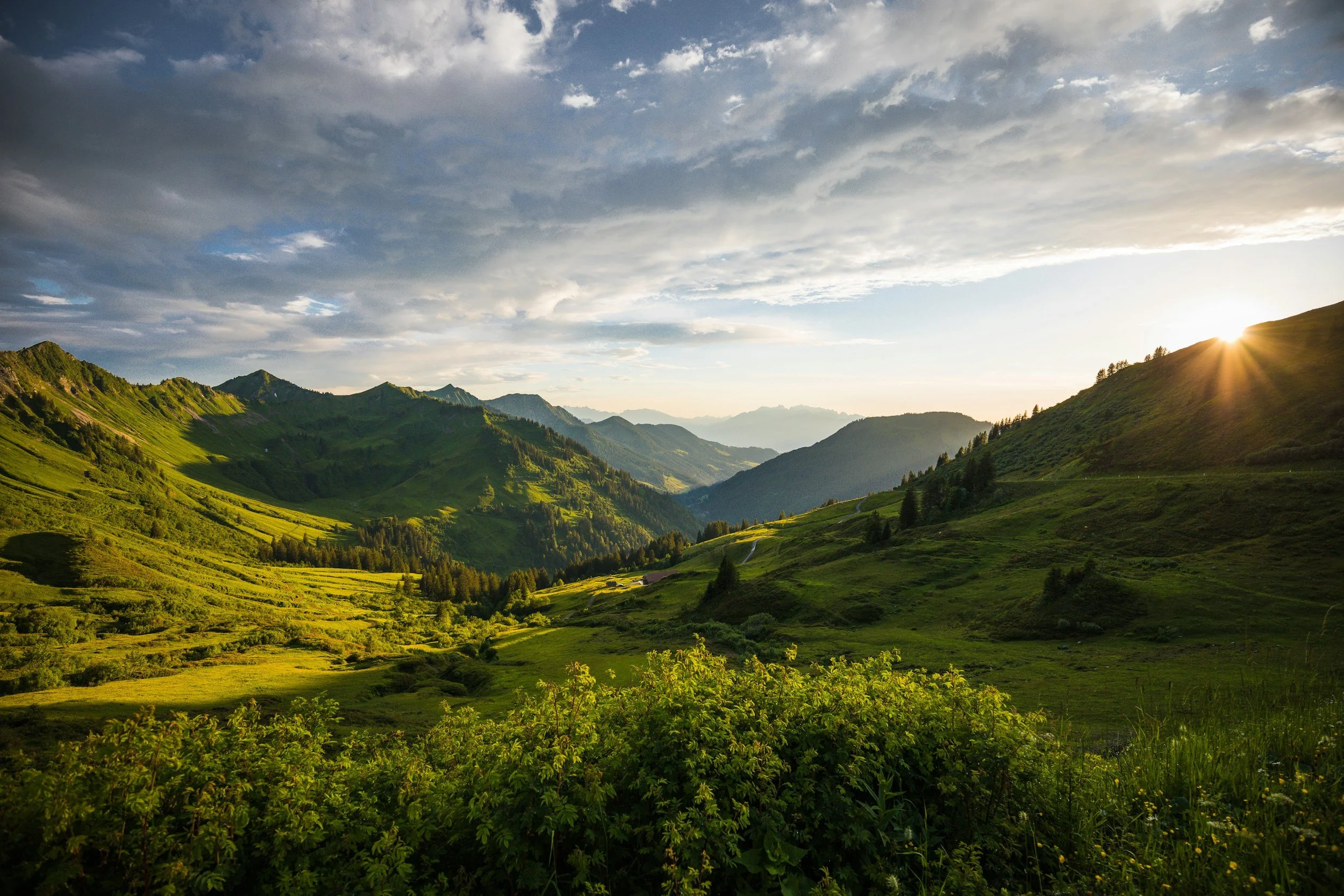 Sunset over green rolling hills and mountains in a valley with scattered trees and a partly cloudy sky.