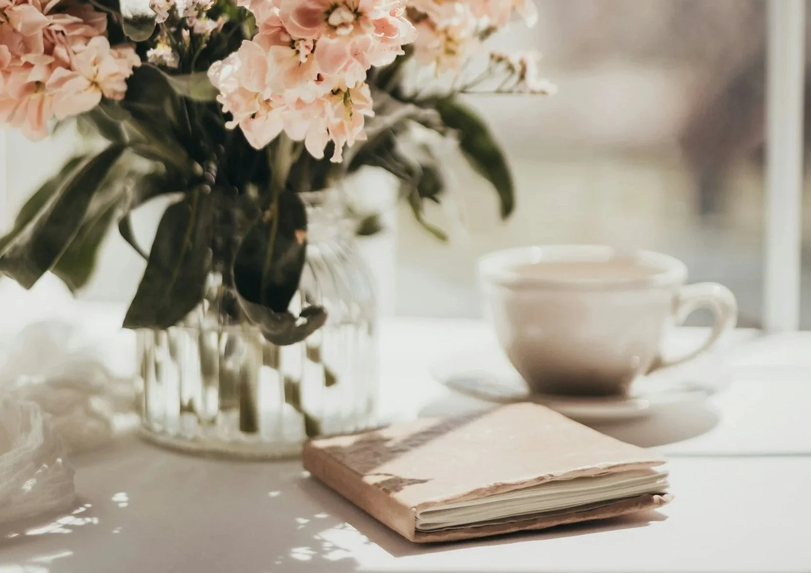 A vase with pink and white flowers, a closed notebook, a teacup with a saucer, all placed on a white surface with natural light.