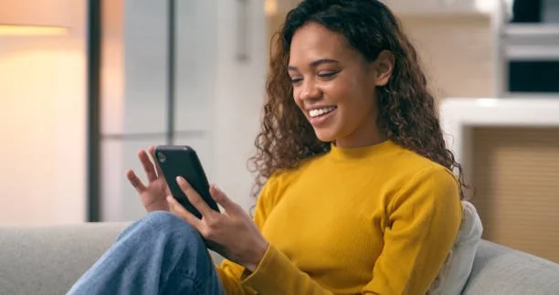 Woman in yellow sweater sitting on couch looking at her phone and smiling