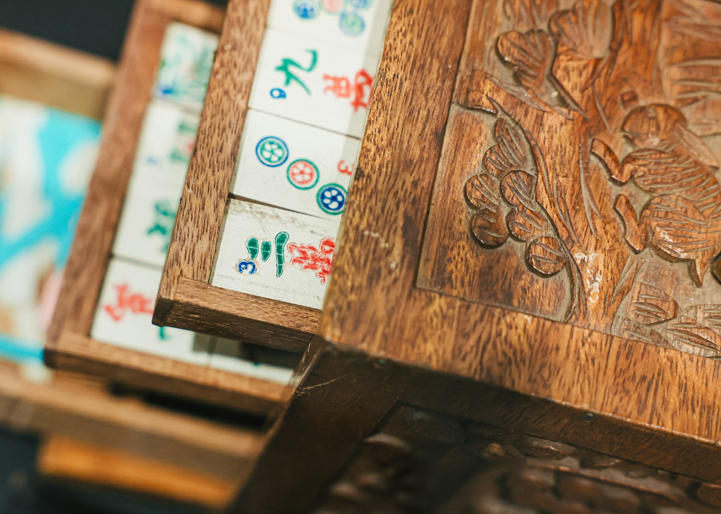 Close-up of a wooden game board with intricate carved floral design on the right and a row of mahjong (mah jongg) tiles with colored characters and symbols on the left.