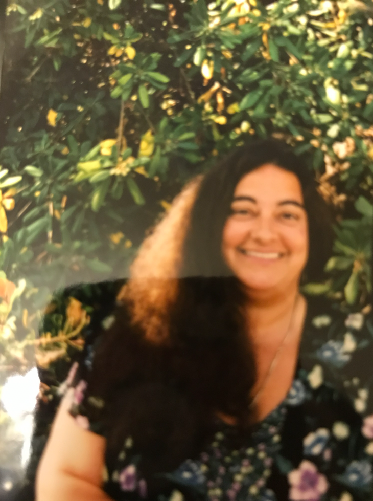 A smiling woman with black hair, wearing a floral dress, standing in front of green leafy bushes with yellow flowers. Private American Mah Jongg Lesson Teacher, Lori Brody.
