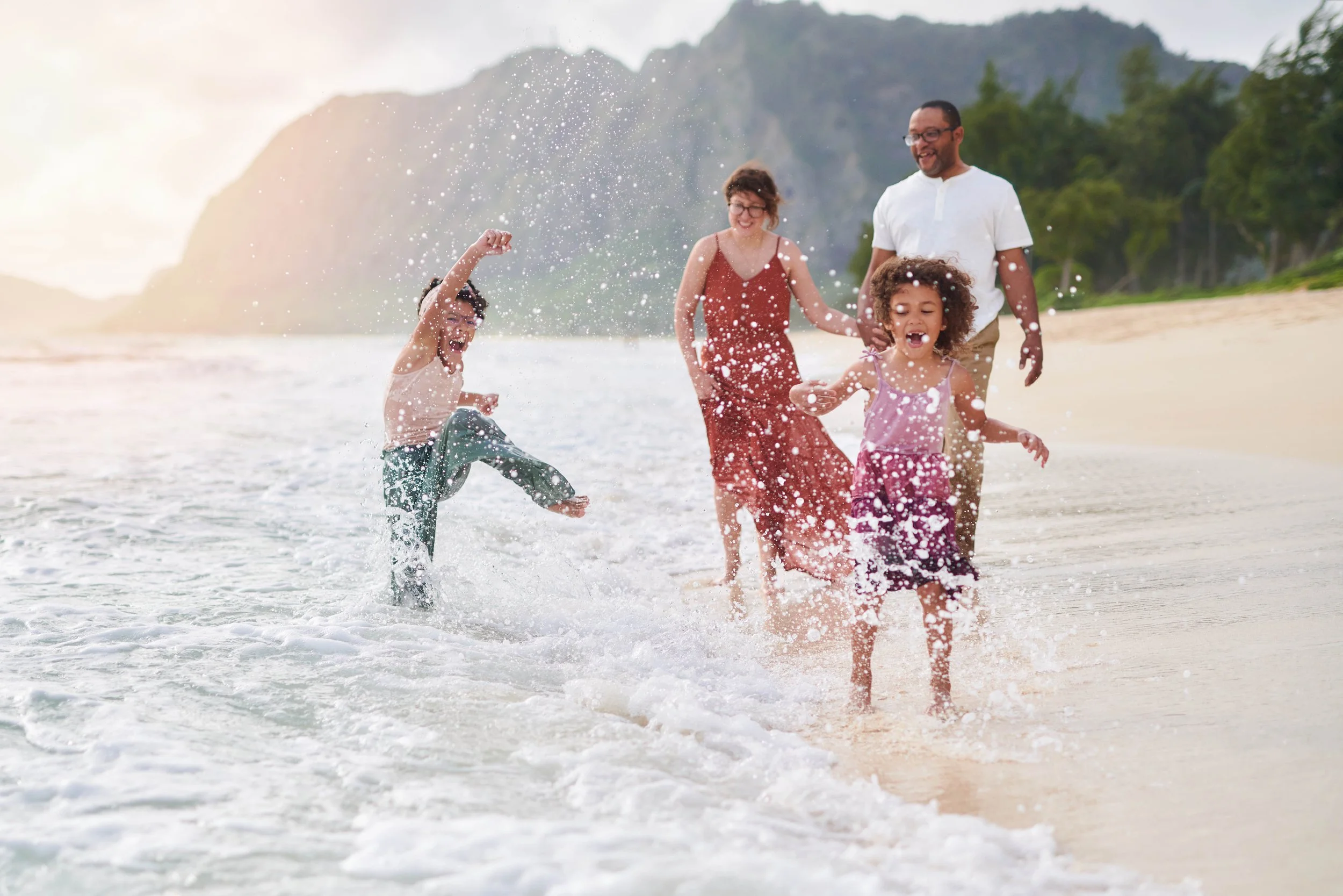 A family of four playing and running along the shoreline at the beach, splashing water, with mountains and trees in the background.