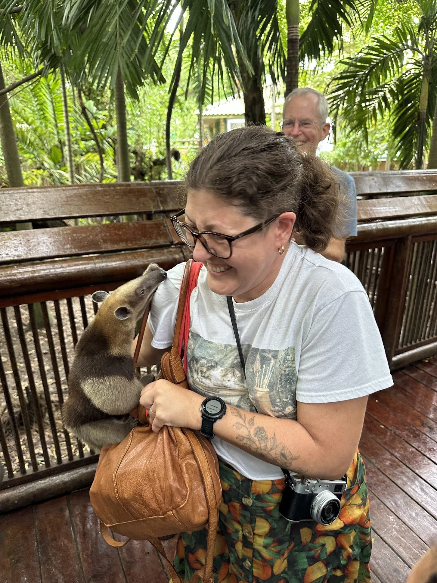 Woman holding a ferret while smiling, with a man sitting on a wooden bench in the background in a lush, green outdoor setting.