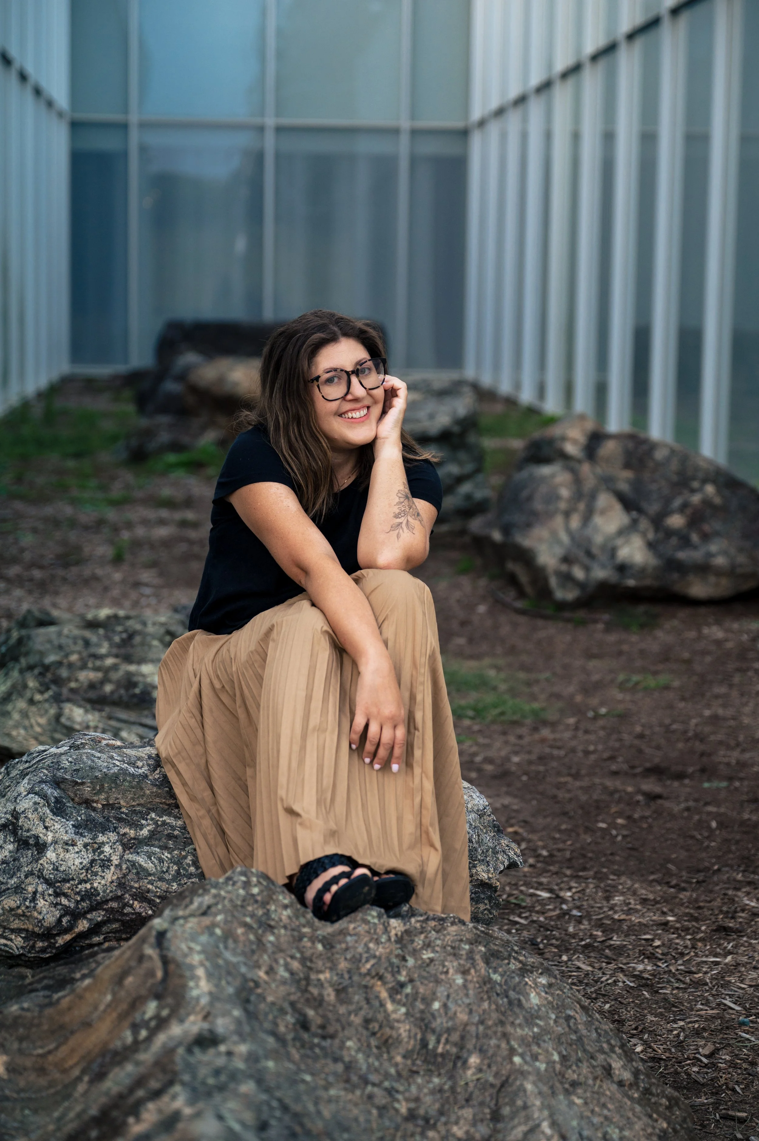 A woman with glasses and long brown hair, dressed in a black shirt and tan pleated skirt, sitting on a rock outdoors, smiling and resting her chin on her hand, with a greenhouse or glass building in the background.