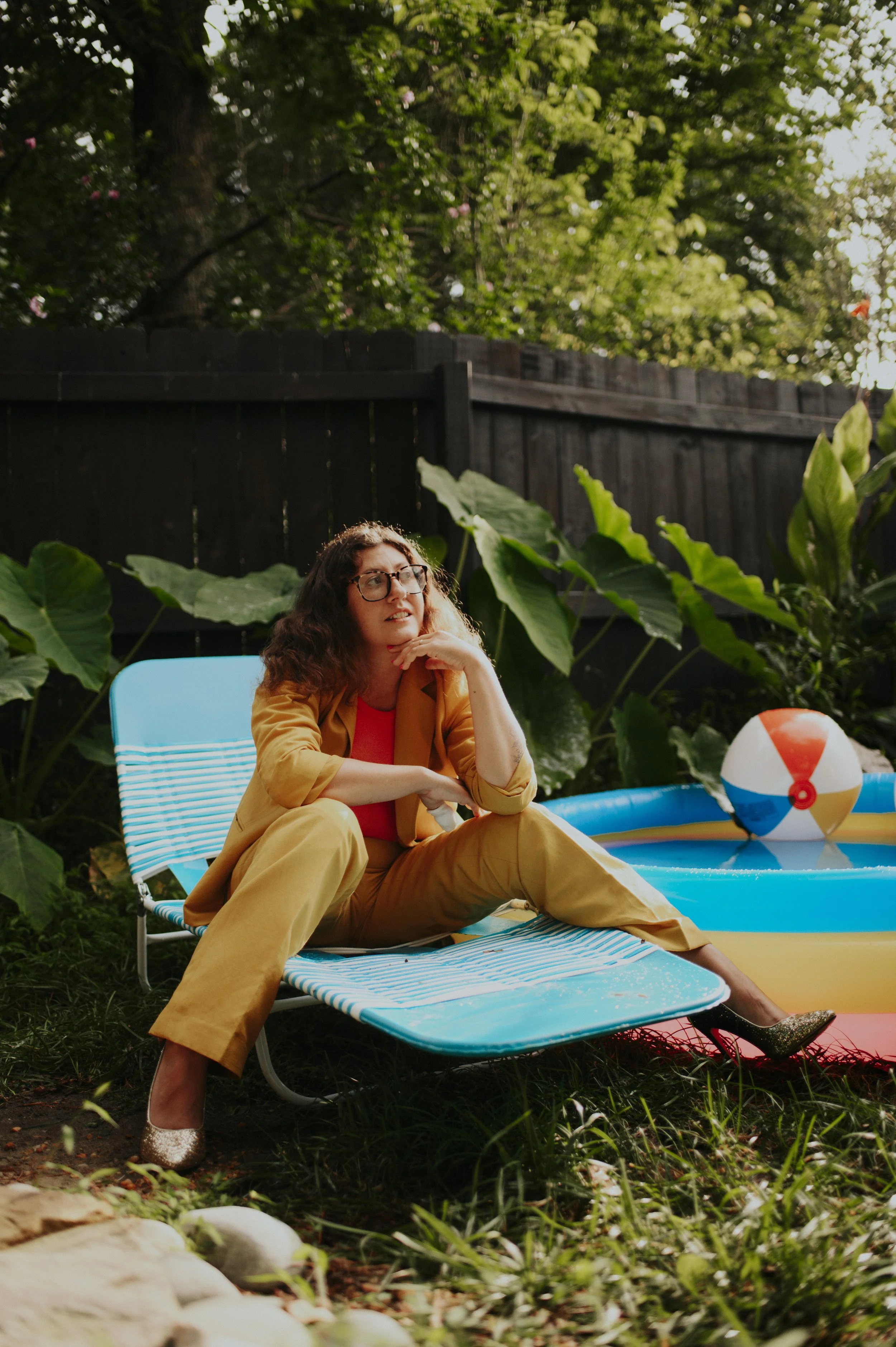 A woman with curly hair and glasses is sitting on a blue and white striped lounge chair outdoors, resting her chin on her hand, with an inflatable pool and beach ball nearby, surrounded by green plants and a wooden fence.