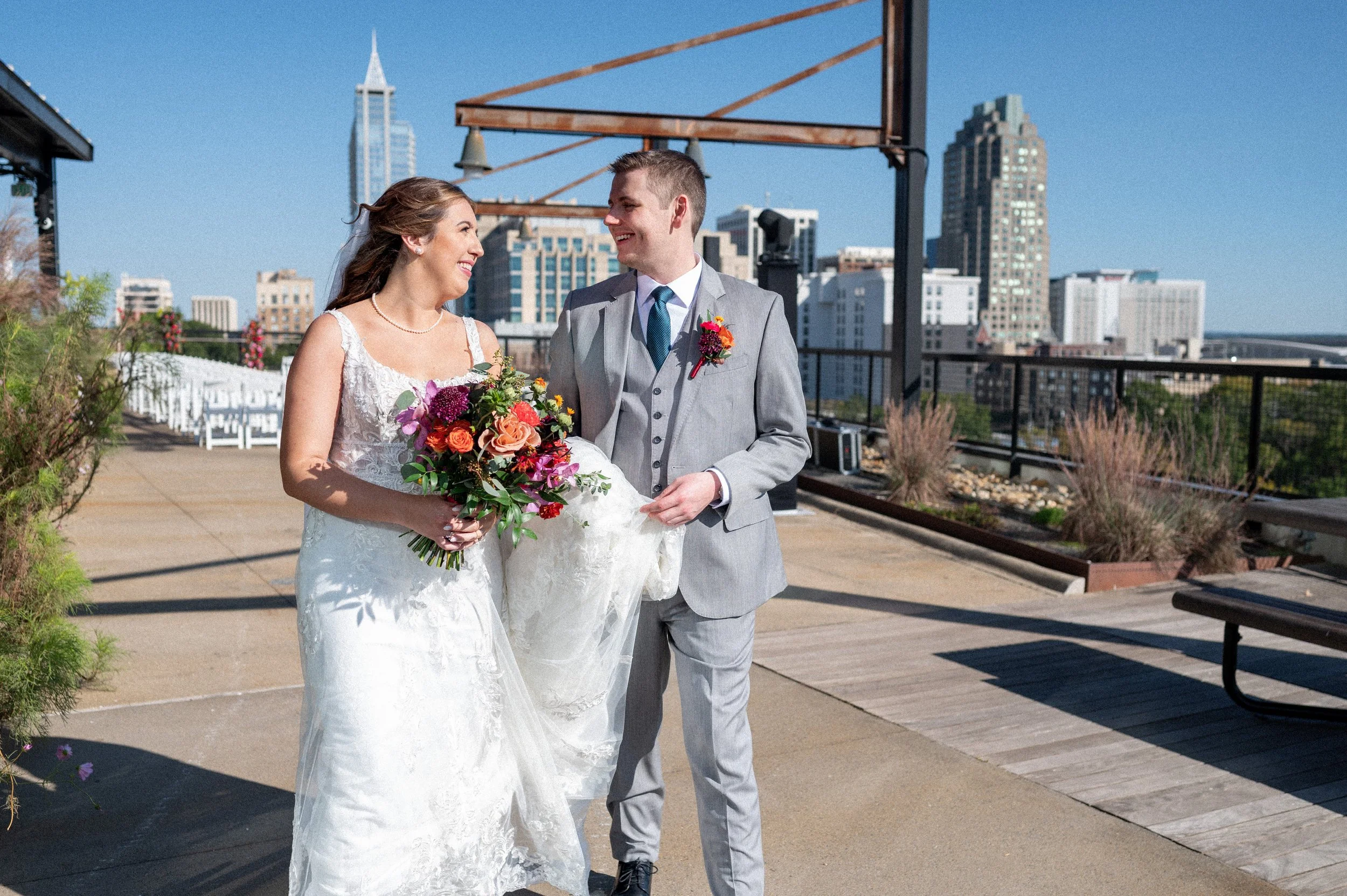 A bride and groom smiling and looking at each other on a rooftop terrace with city skyline in the background, the bride holding a colorful bouquet.