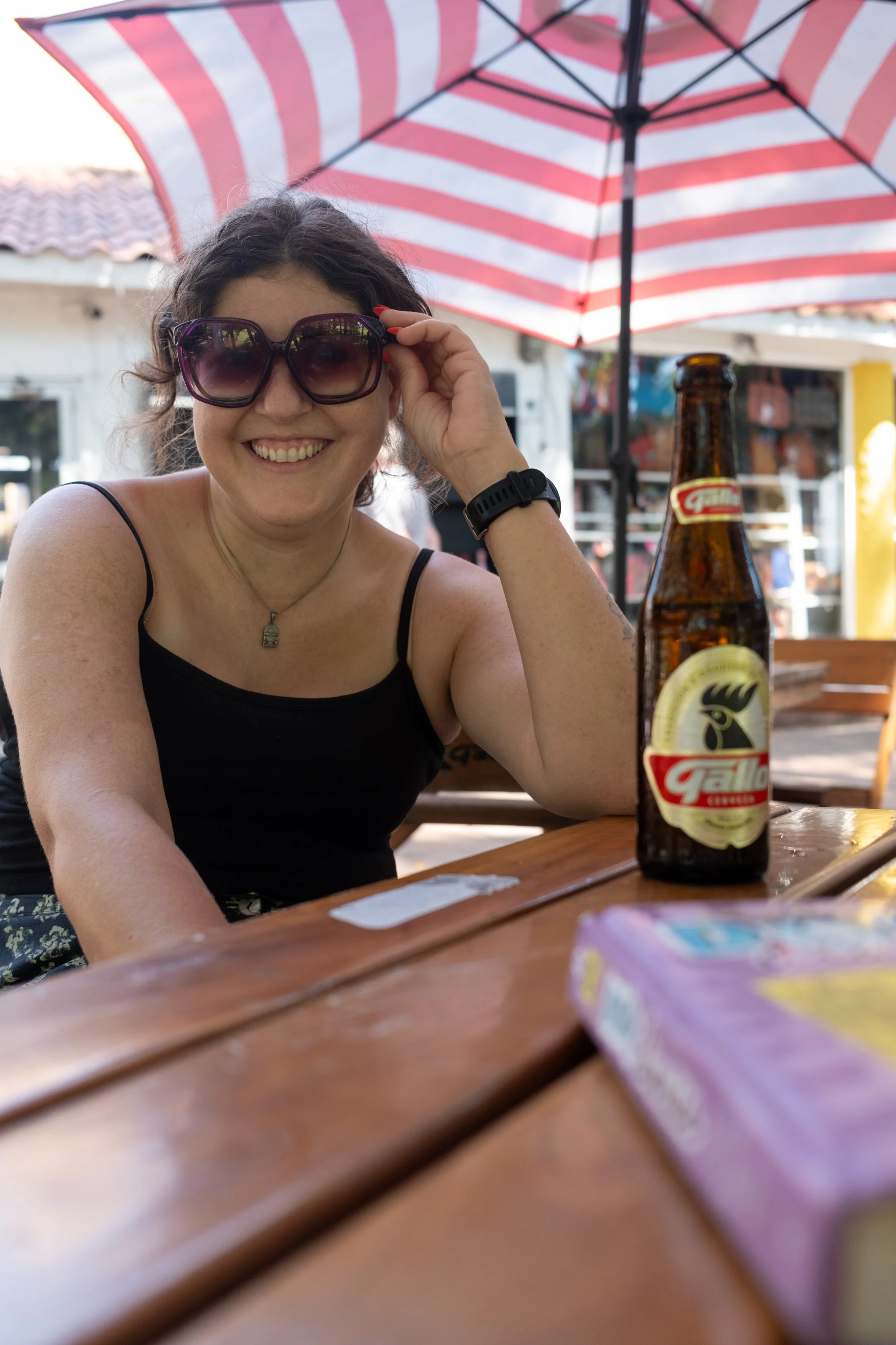 A woman smiling and wearing sunglasses sitting at an outdoor table under a red and white striped umbrella, with a bottle of Gallo beer on the table.