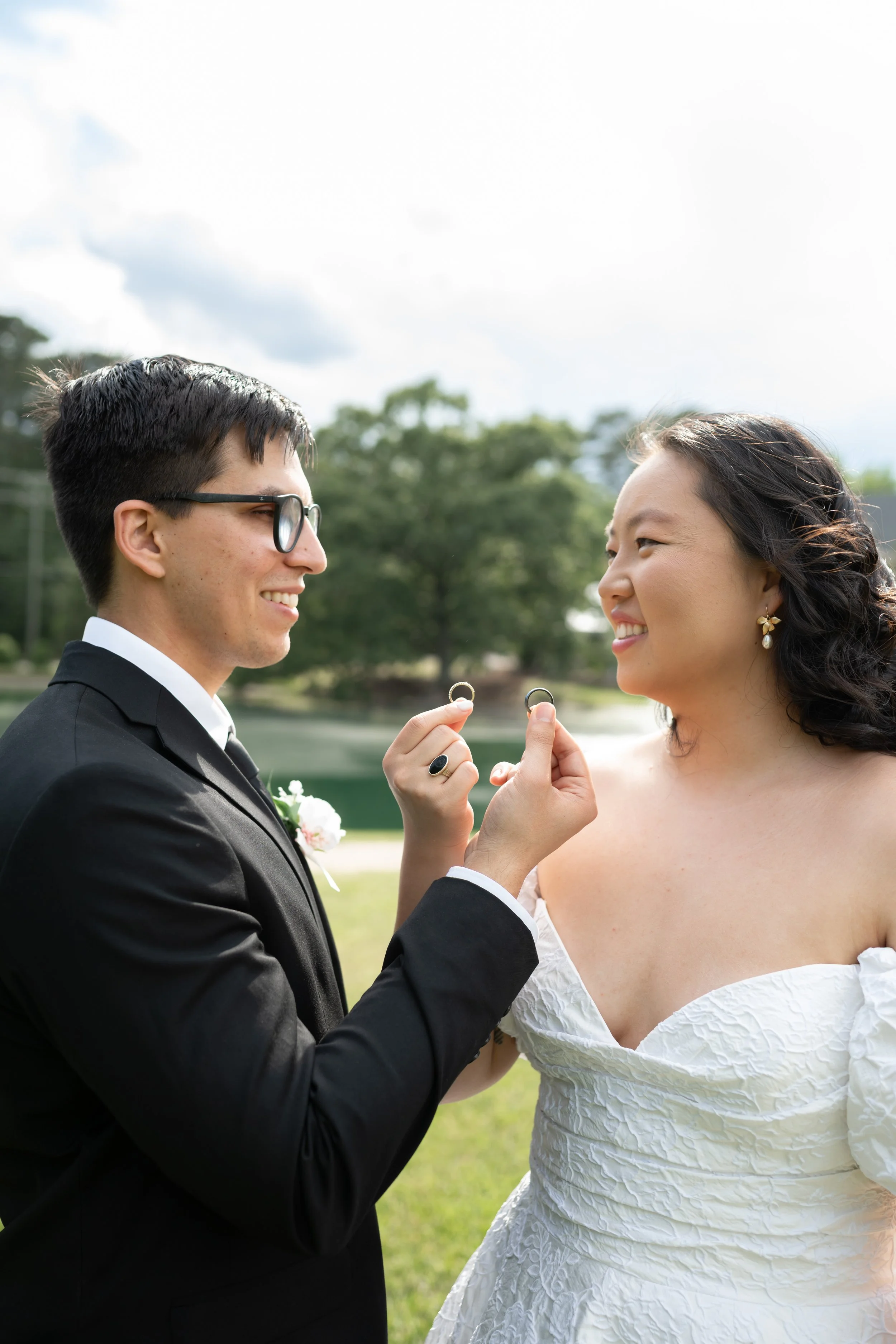 A couple gets married outside holding wedding rings, with a lake and trees in the background.