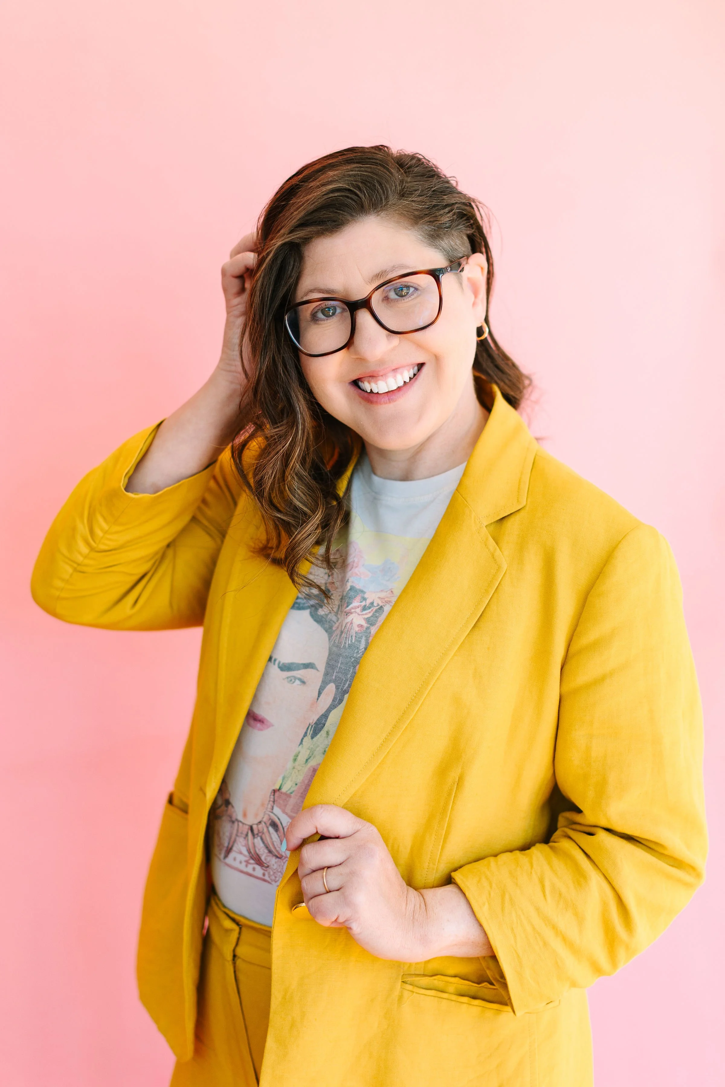 A woman with glasses wearing a yellow blazer over a graphic T-shirt, smiling and touching her hair against a pink background.