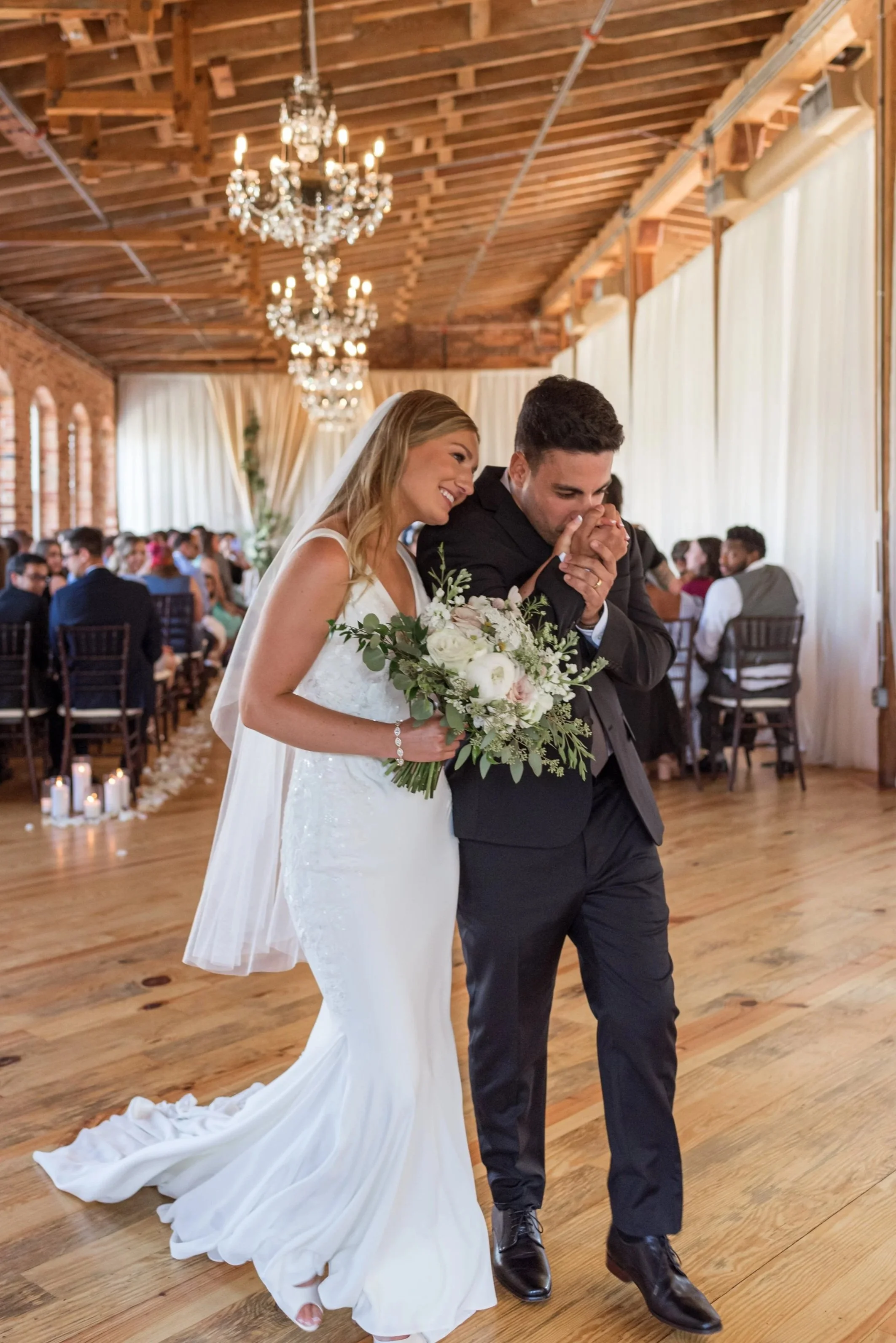 A bride and groom share a kiss during their wedding ceremony in a rustic venue with wooden floors and chandeliers.