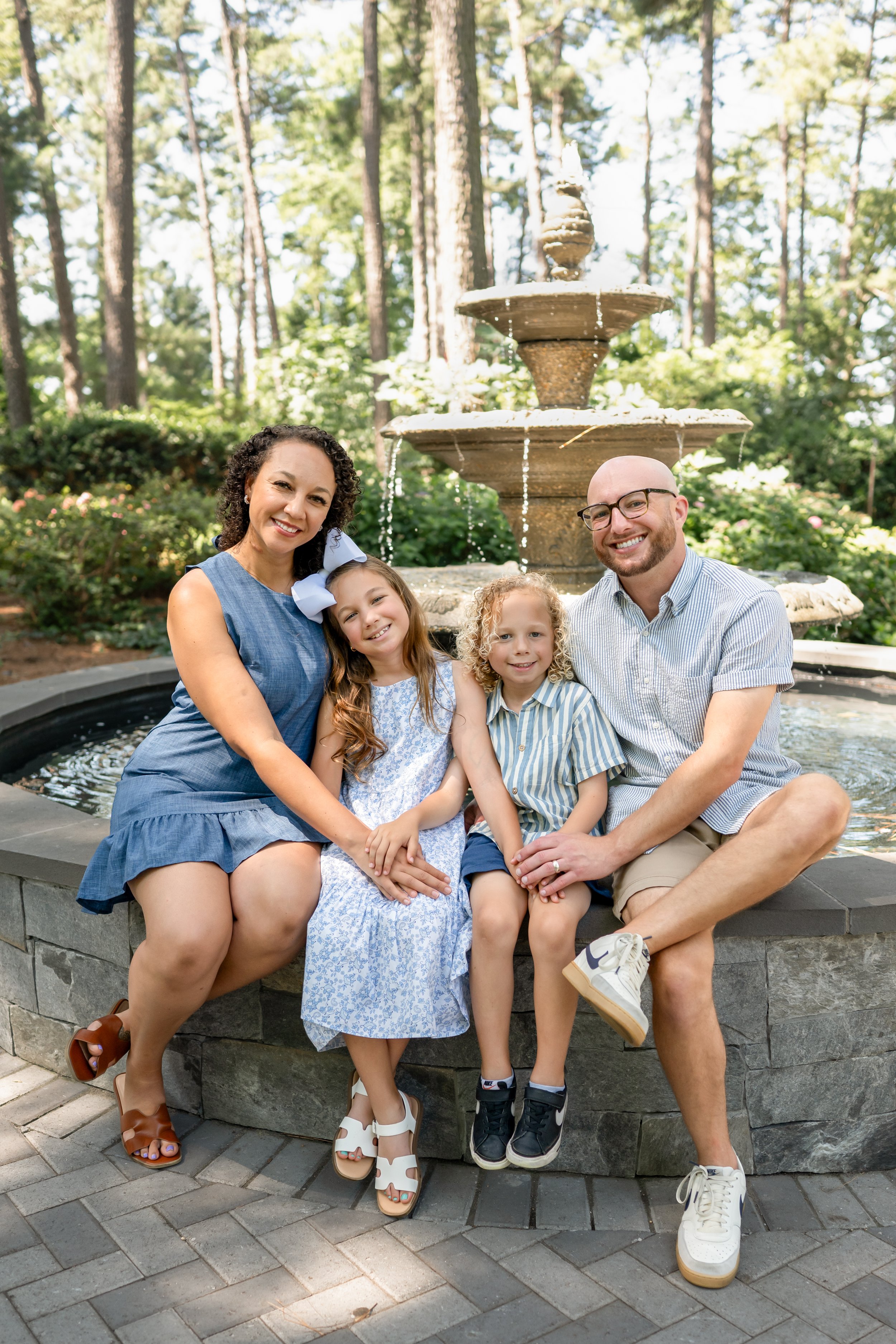 Family of four sitting on a stone fountain in a lush green park during daytime. The mother and father are smiling, with two children, a girl and a boy, wearing summer clothes.