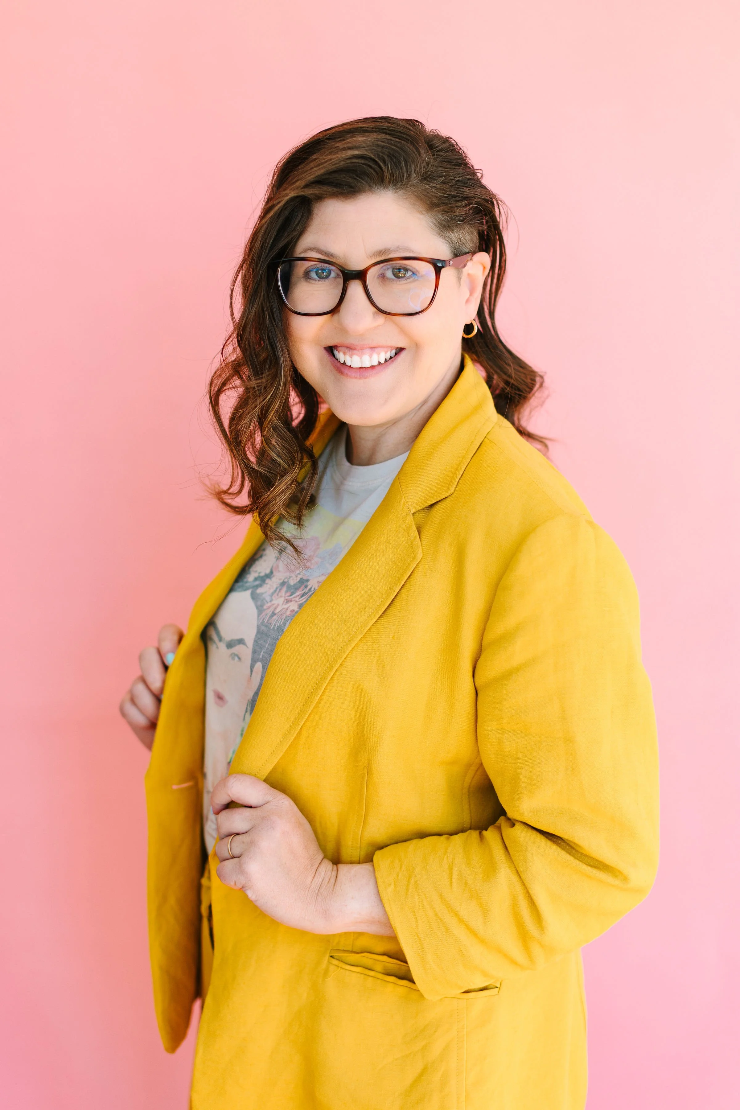 A woman wearing glasses and a yellow blazer, smiling, holding the blazer lapel with a pink background.