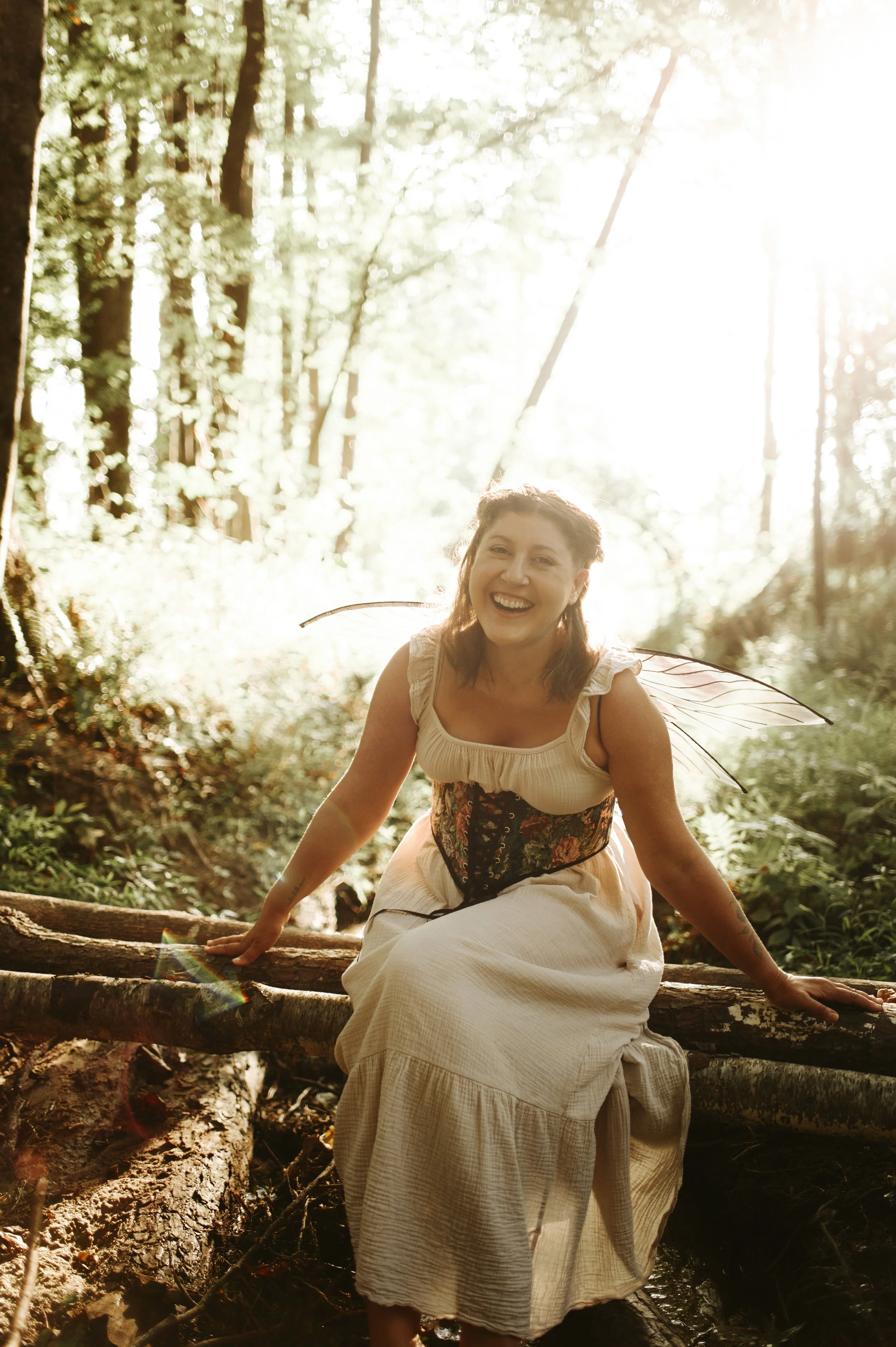 A woman with fairy wings and a colorful corset smiling and sitting on a fallen log in a sunlit wooded forest.