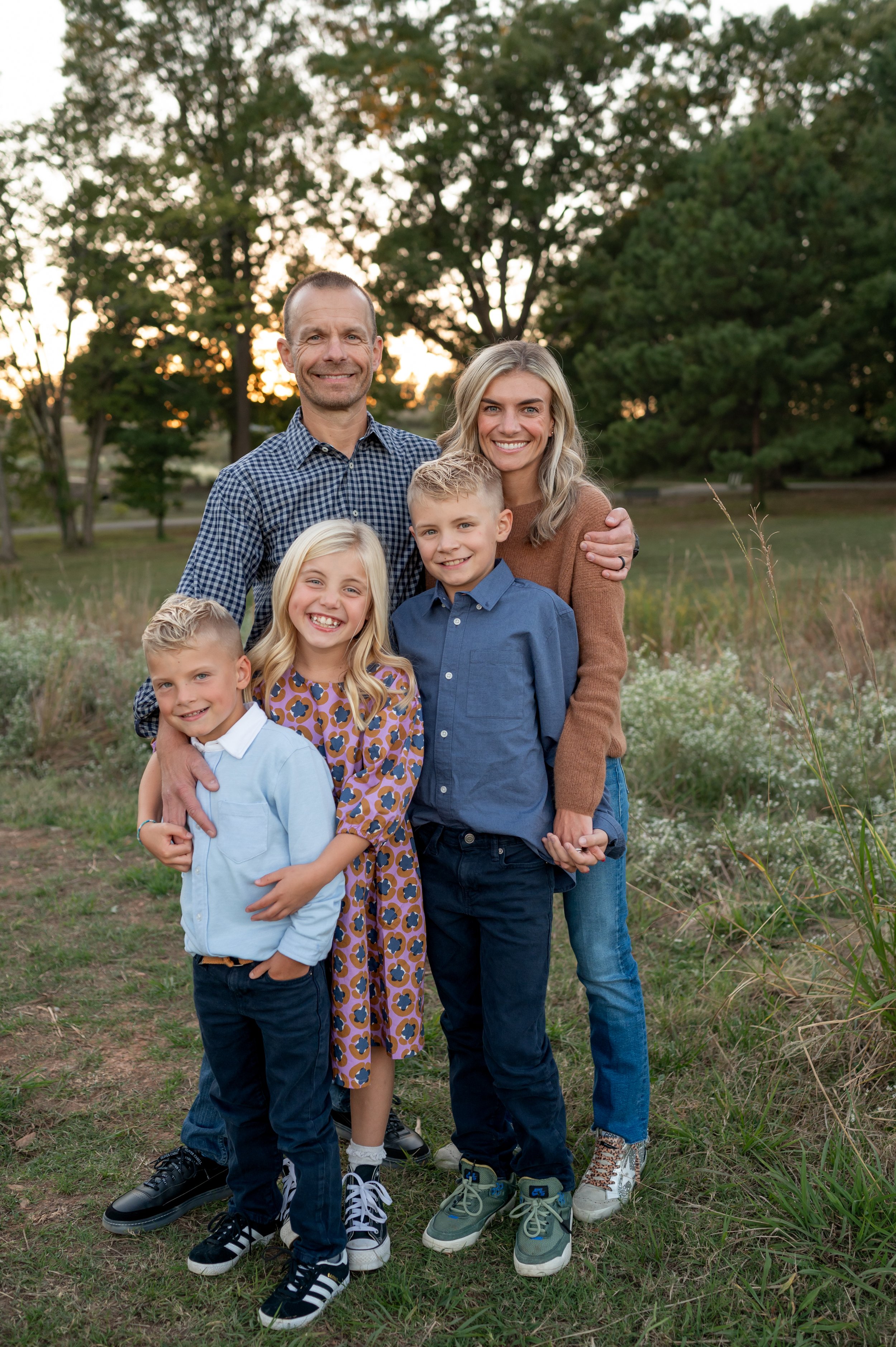 A family of six standing outdoors in a park at sunset, smiling and embracing each other.