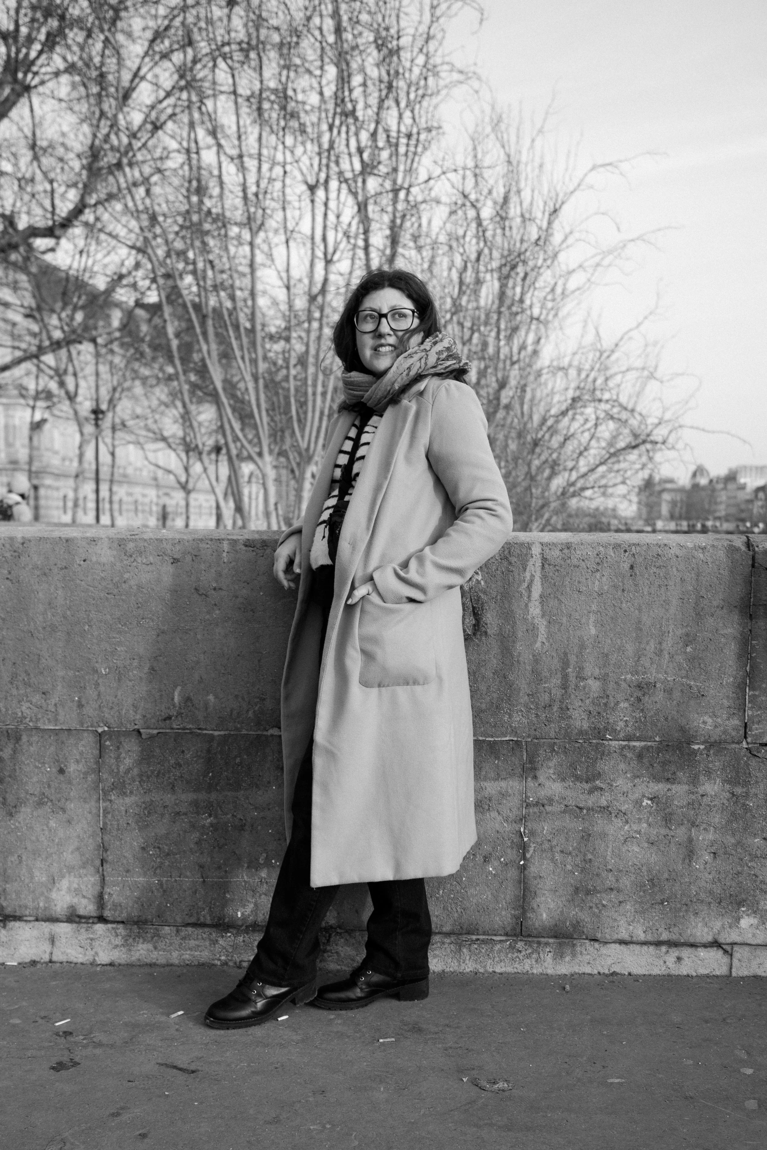 A woman with glasses, wearing a long coat and scarf, stands outdoors against a stone wall, with trees and city buildings in the background.