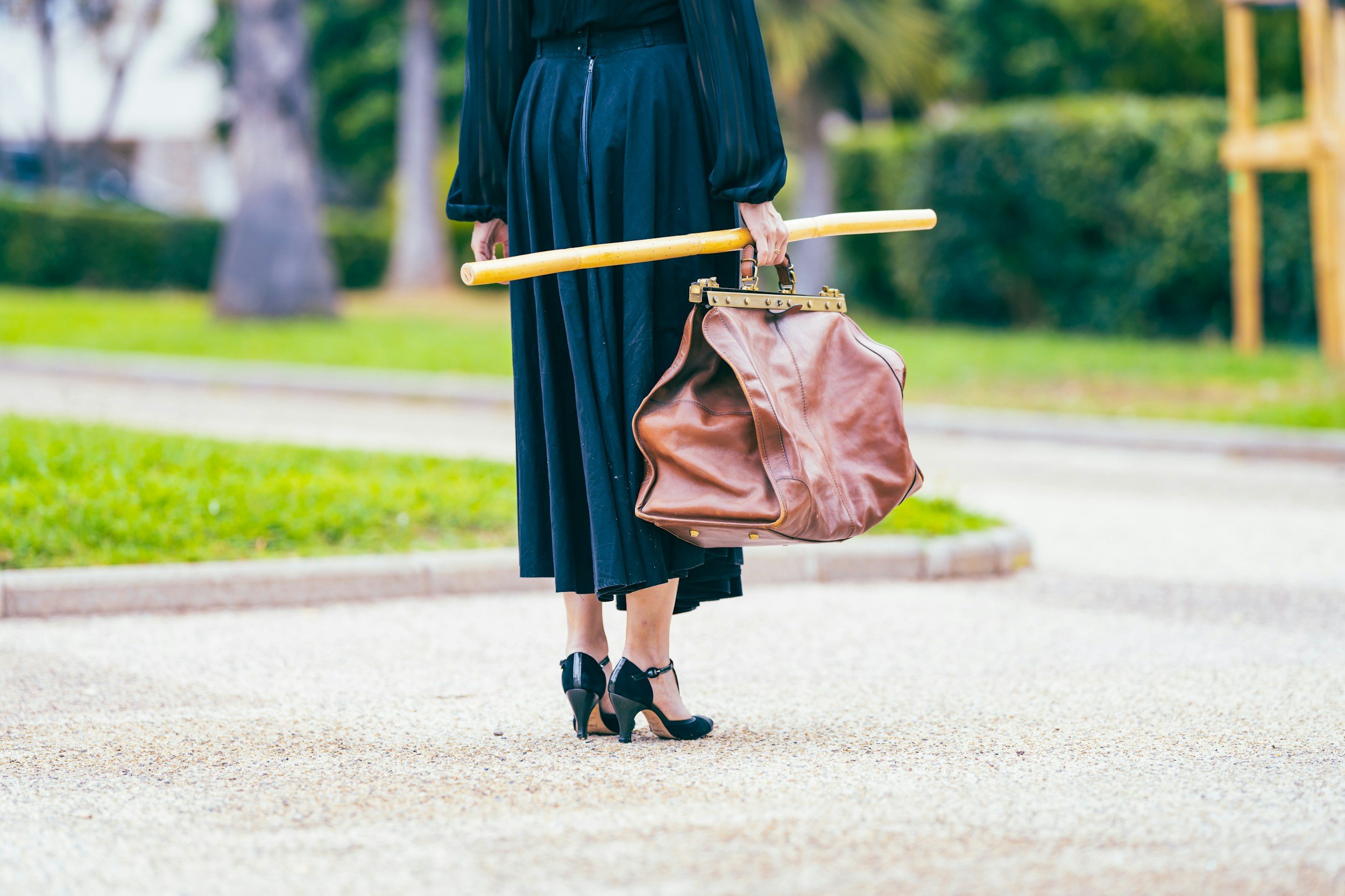 Person dressed as a graduate holding a yellow diploma rod and a brown leather handbag, standing outdoors on a sidewalk with green trees and grass in the background.