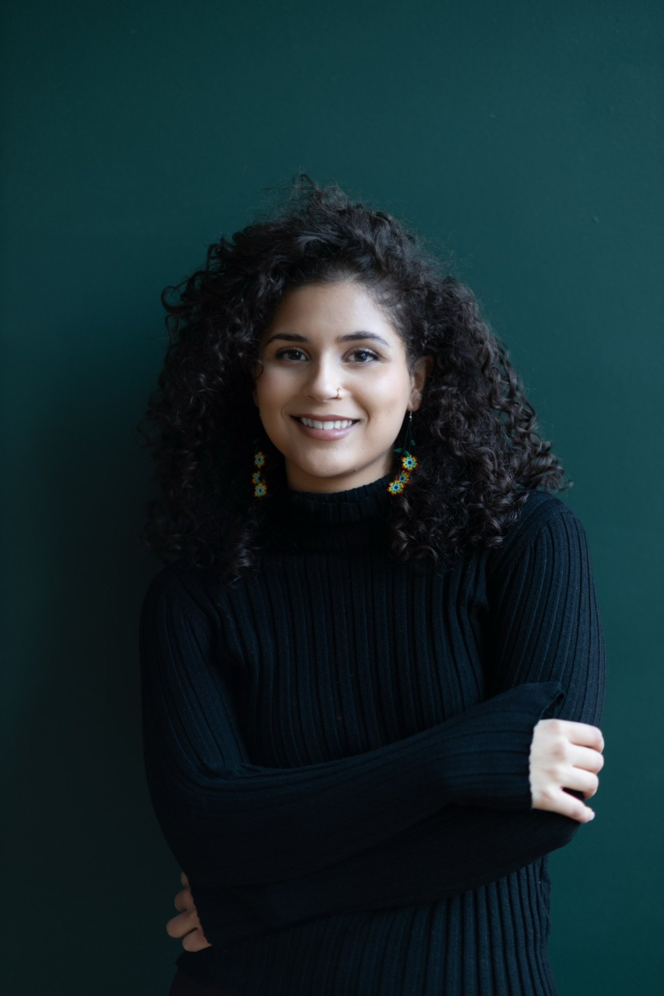 A young woman with dark curly hair smiling, wearing a black ribbed sweater and colorful flower earrings, posing against a dark green background.