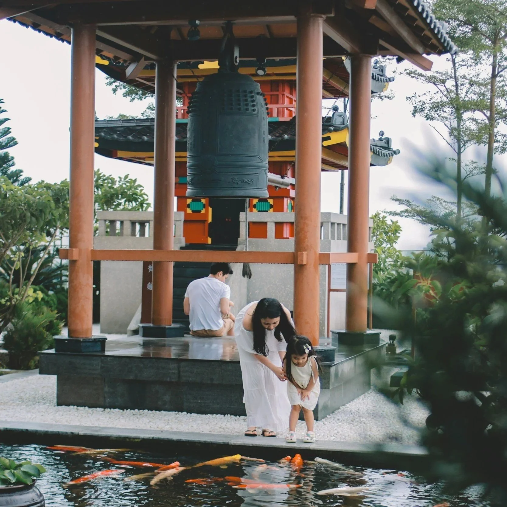 A woman and a young girl feed koi fish in a pond near a traditional Asian-style temple with large bell and wooden pillars.