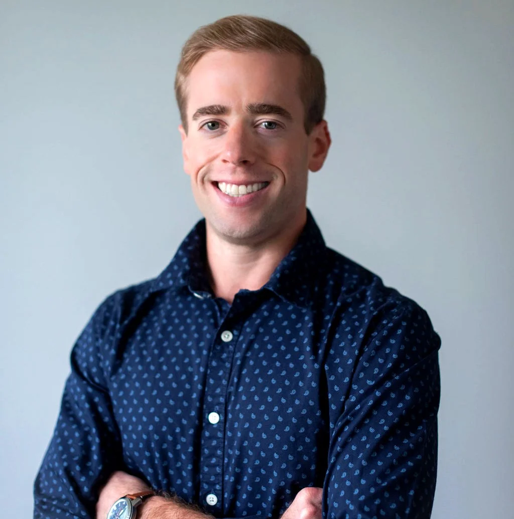 A young man with light brown hair, smiling, wearing a dark blue shirt with small light blue patterns, standing with arms crossed against a plain light gray background.
