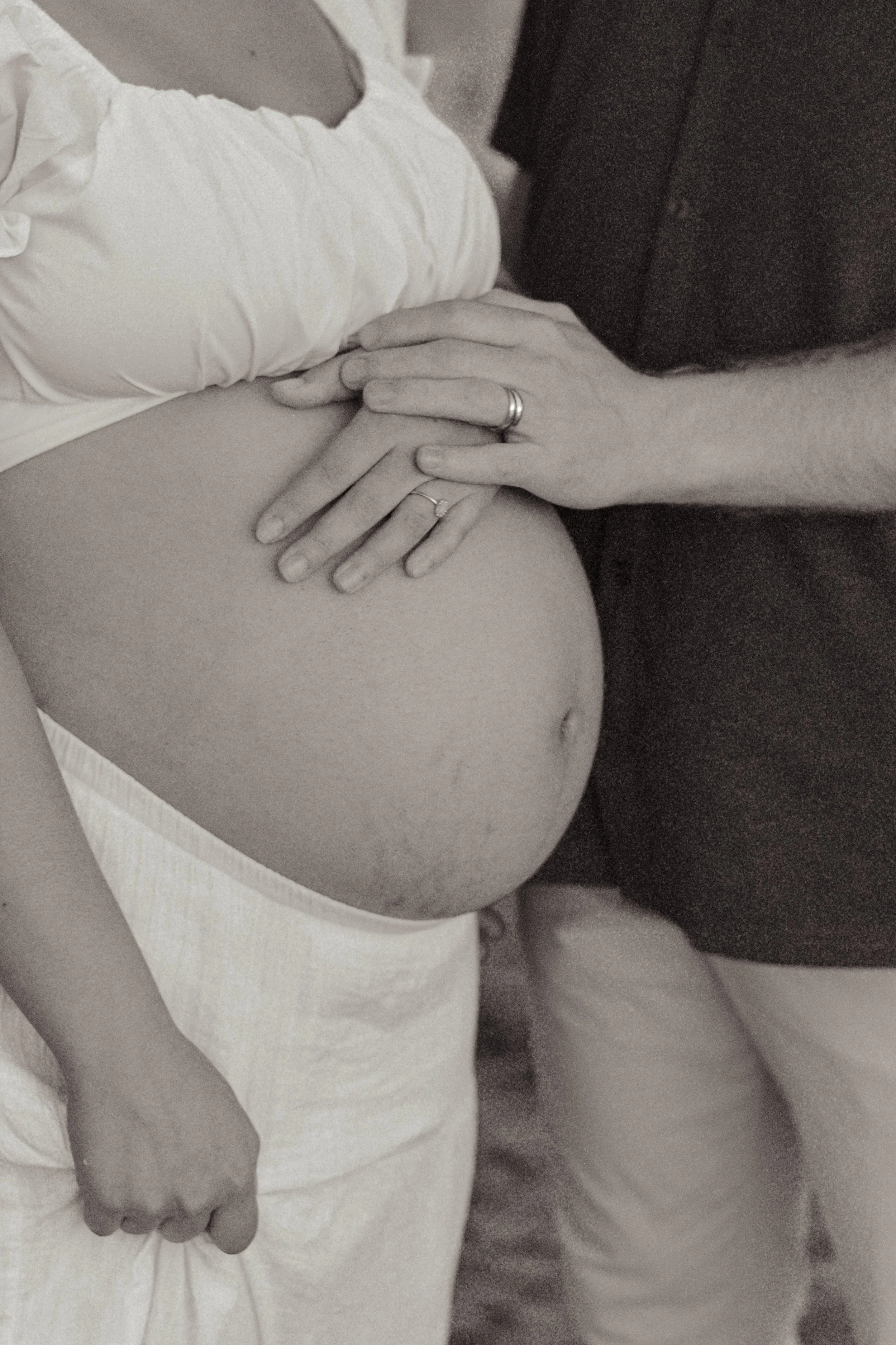 Close-up of a pregnant woman lying in bed with a man resting his hand on her belly, both wearing wedding rings.