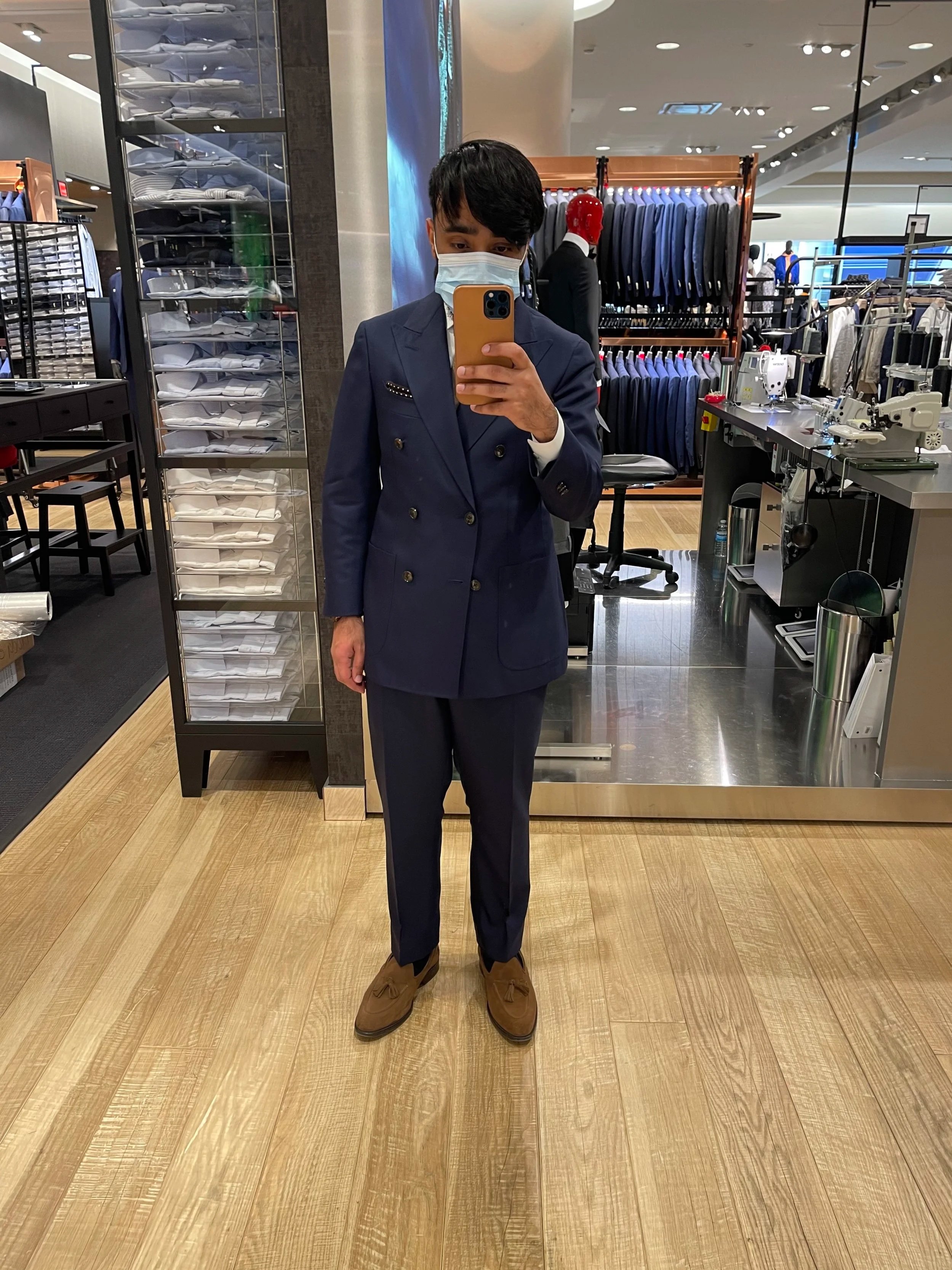 A person wearing a navy blue double-breasted suit with brown loafers, taking a mirror selfie in a clothing store, wearing a face mask.