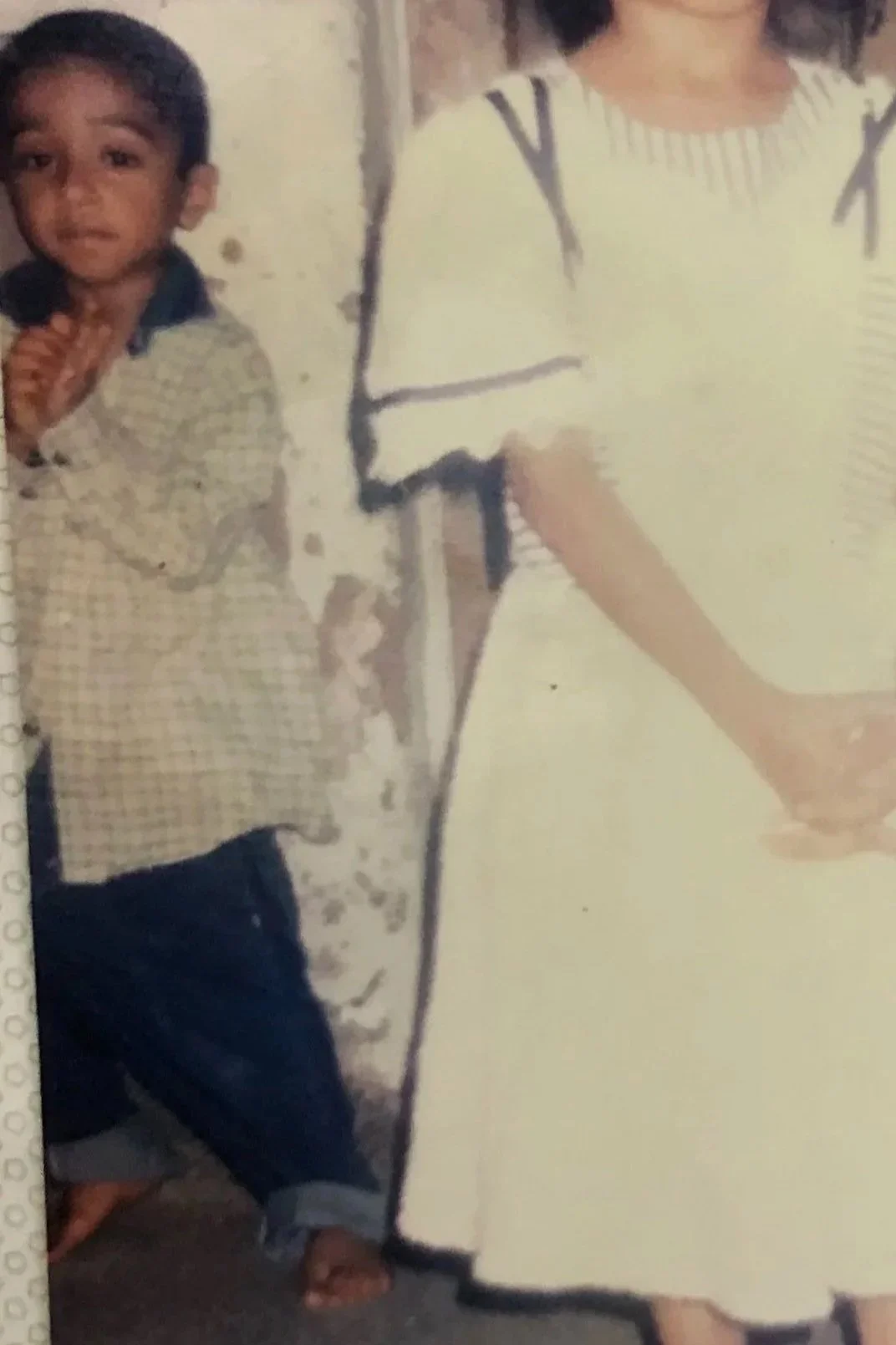 A young boy standing indoors, wearing a patterned shirt and dark pants, with a woman in a yellow and white striped dress next to him, holding a piece of paper, in a room with a plain wall.