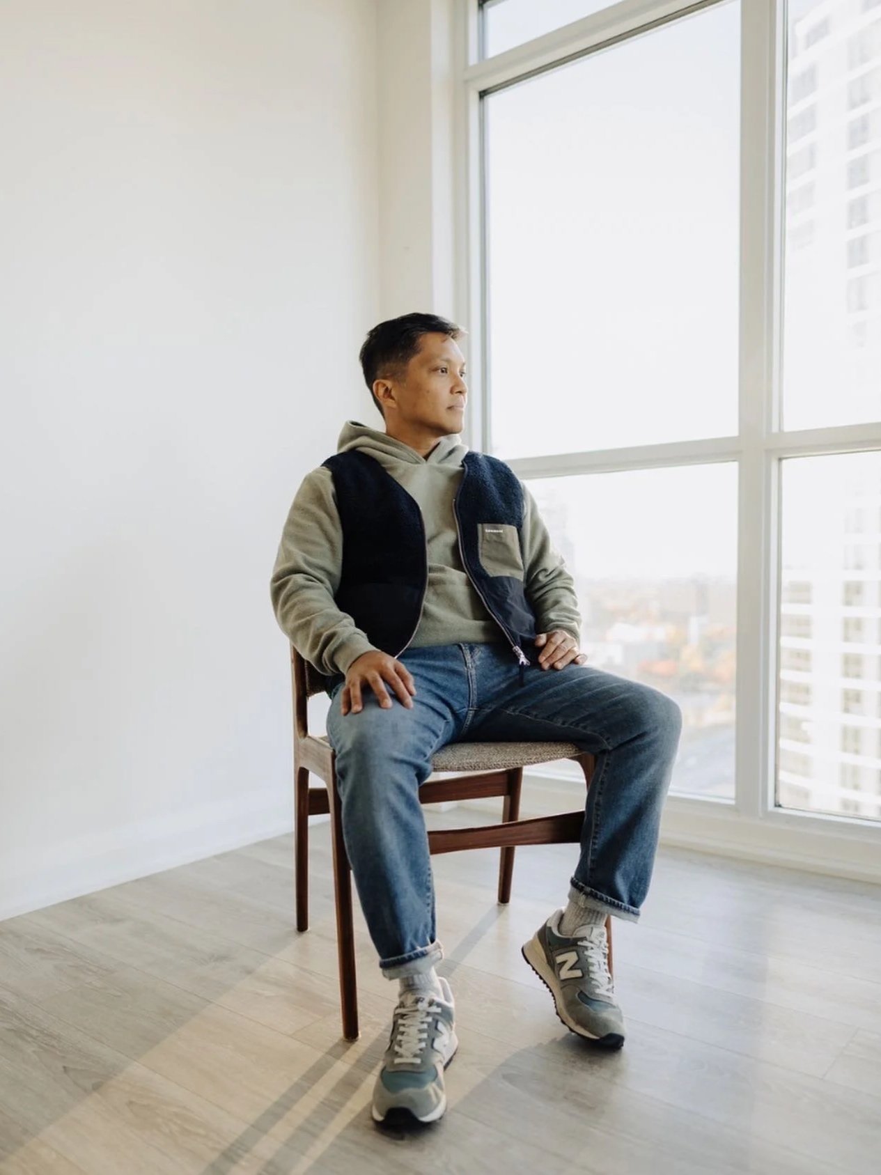 Man sitting on a chair by a large window, looking outside, in a minimalistic room with wooden flooring.
