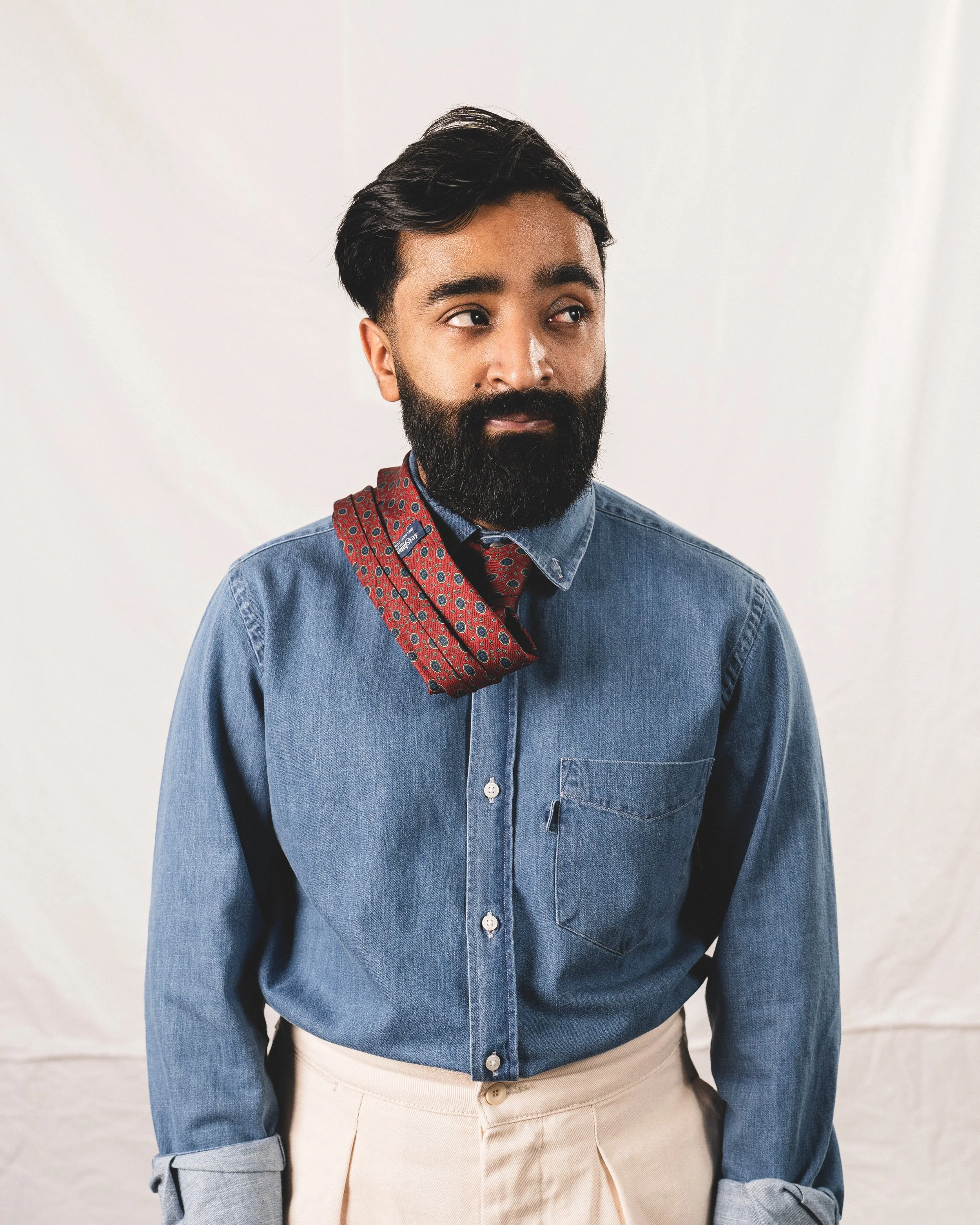 A man with dark hair, beard, and mustache wearing a denim shirt with beige pants, a red patterned neck scarf, and posing against a white background.