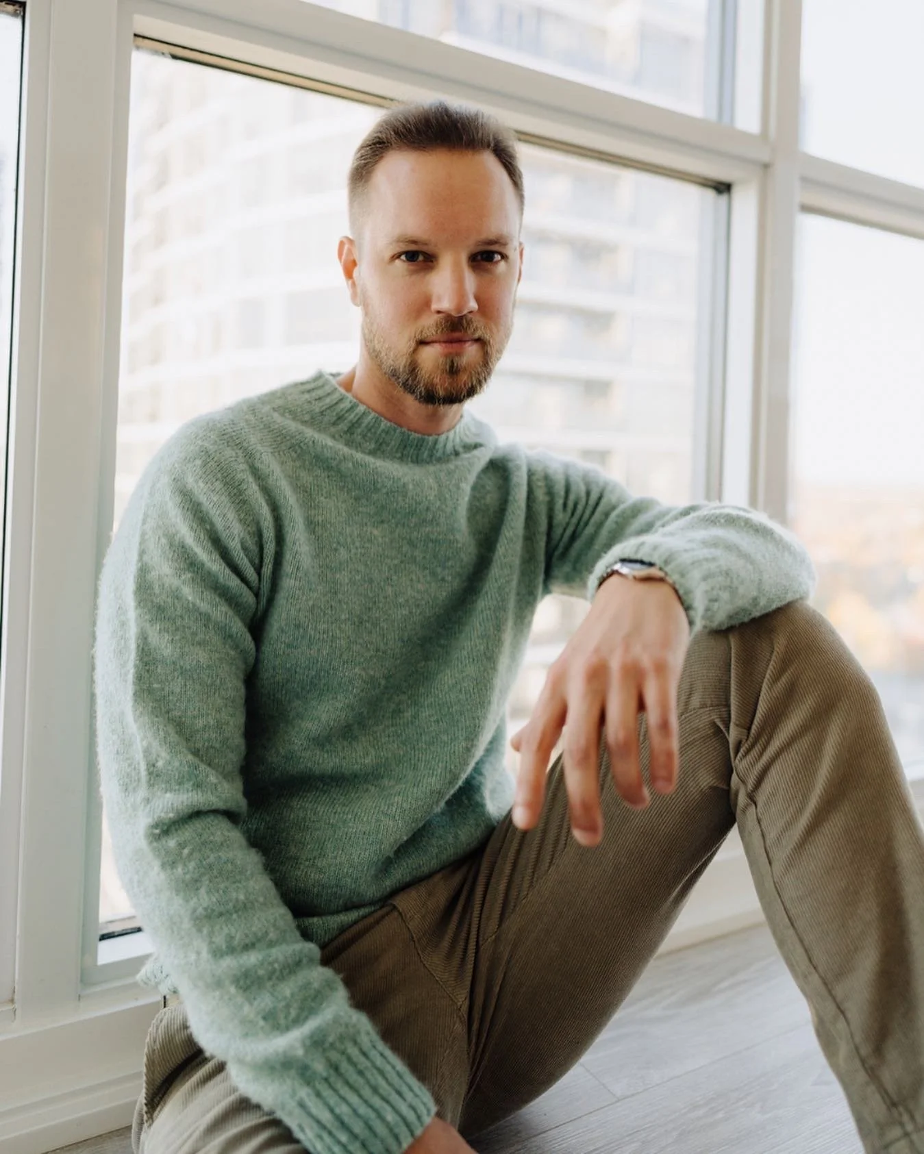 A man with short brown hair, a beard, wearing a light green sweater and beige pants, sitting on the floor by a large window with a cityscape in the background.