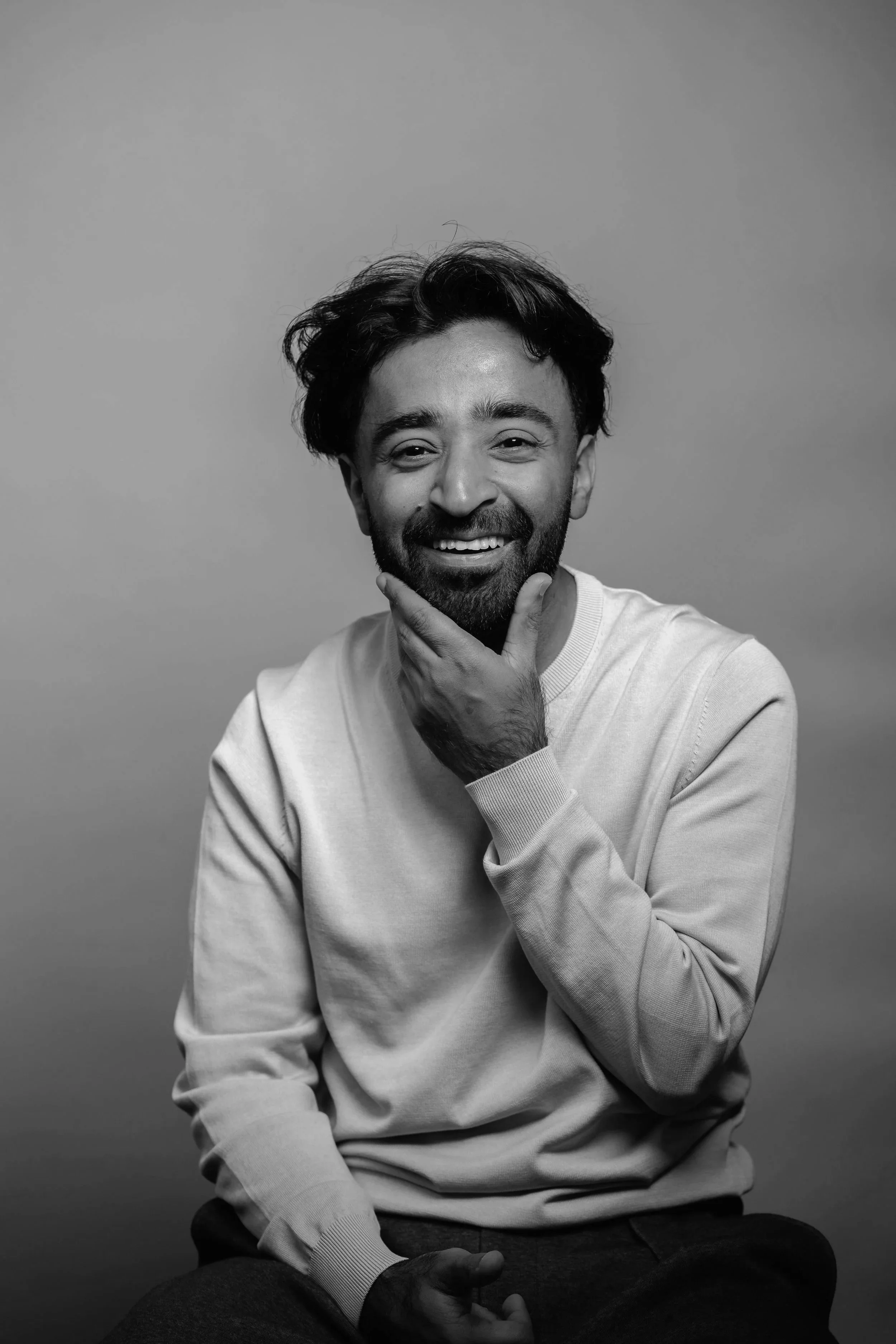 Black-and-white portrait of a smiling man with a beard, short hair, wearing a light-colored sweatshirt, sitting and touching his chin with his hand.