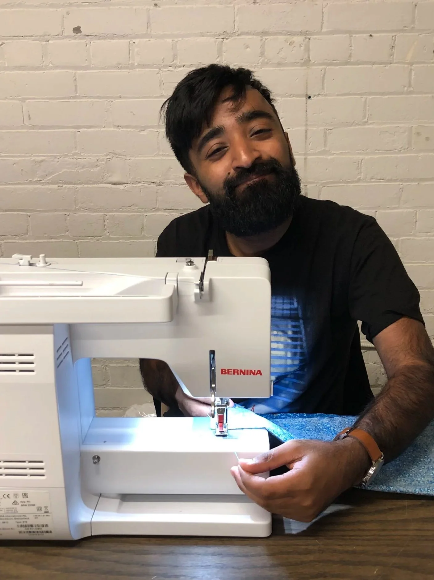 A man with a beard and dark hair, wearing a black t-shirt, sitting at a table using a white sewing machine.