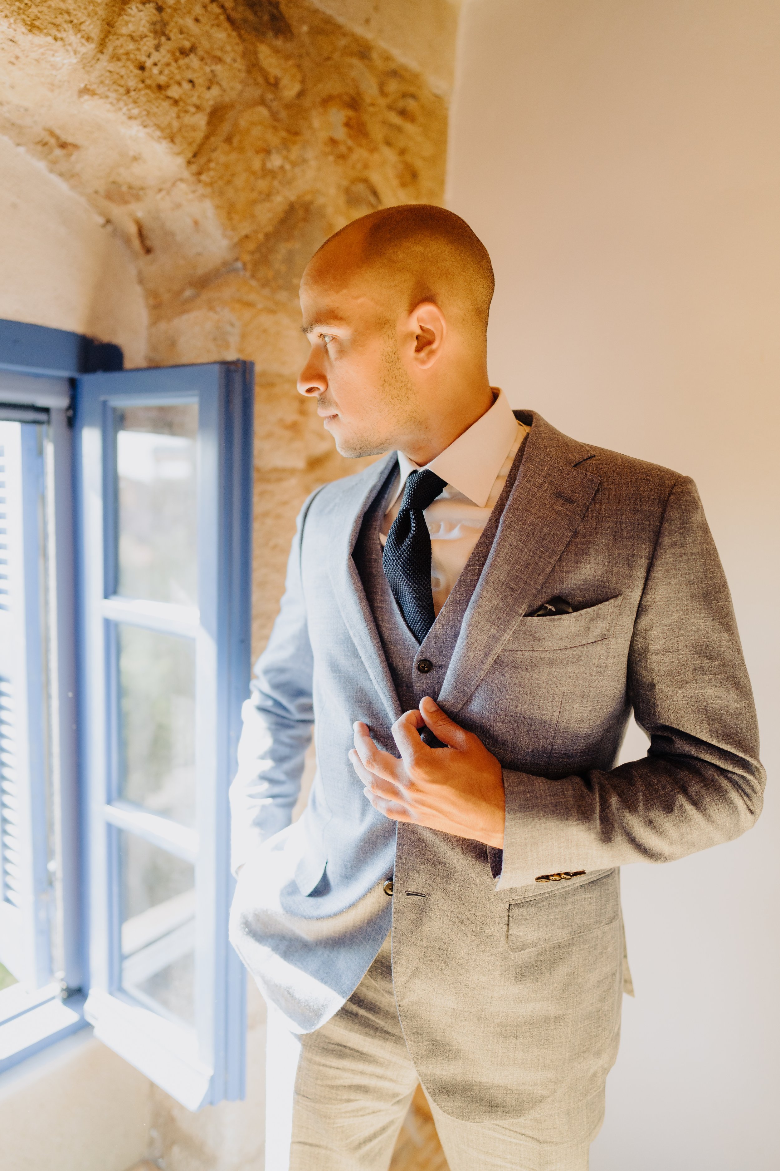 A man in a gray suit looking out of an open window, standing in a room with stone and white walls.