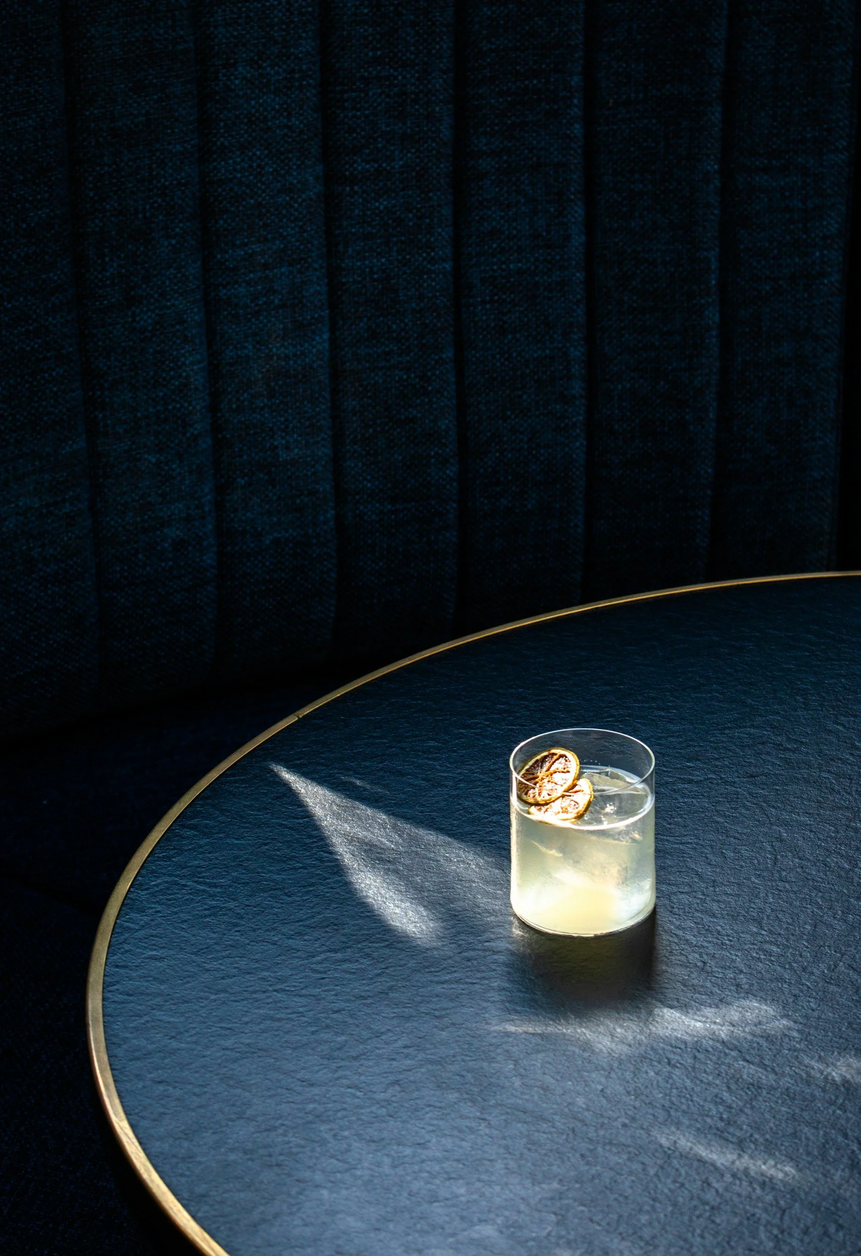 A glass of a light-colored beverage with citrus slices on top, placed on a dark table with a gold rim. The background features a dark, textured upholstered surface.