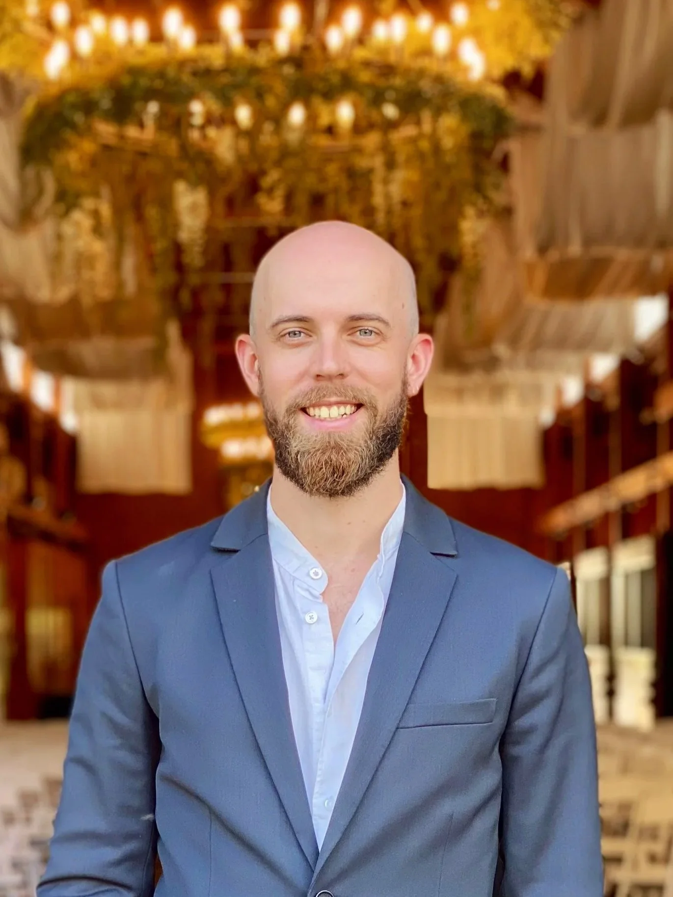 A man with a beard wearing a blue blazer and white shirt, smiling, standing indoors with a wooden interior and hanging lights in the background.