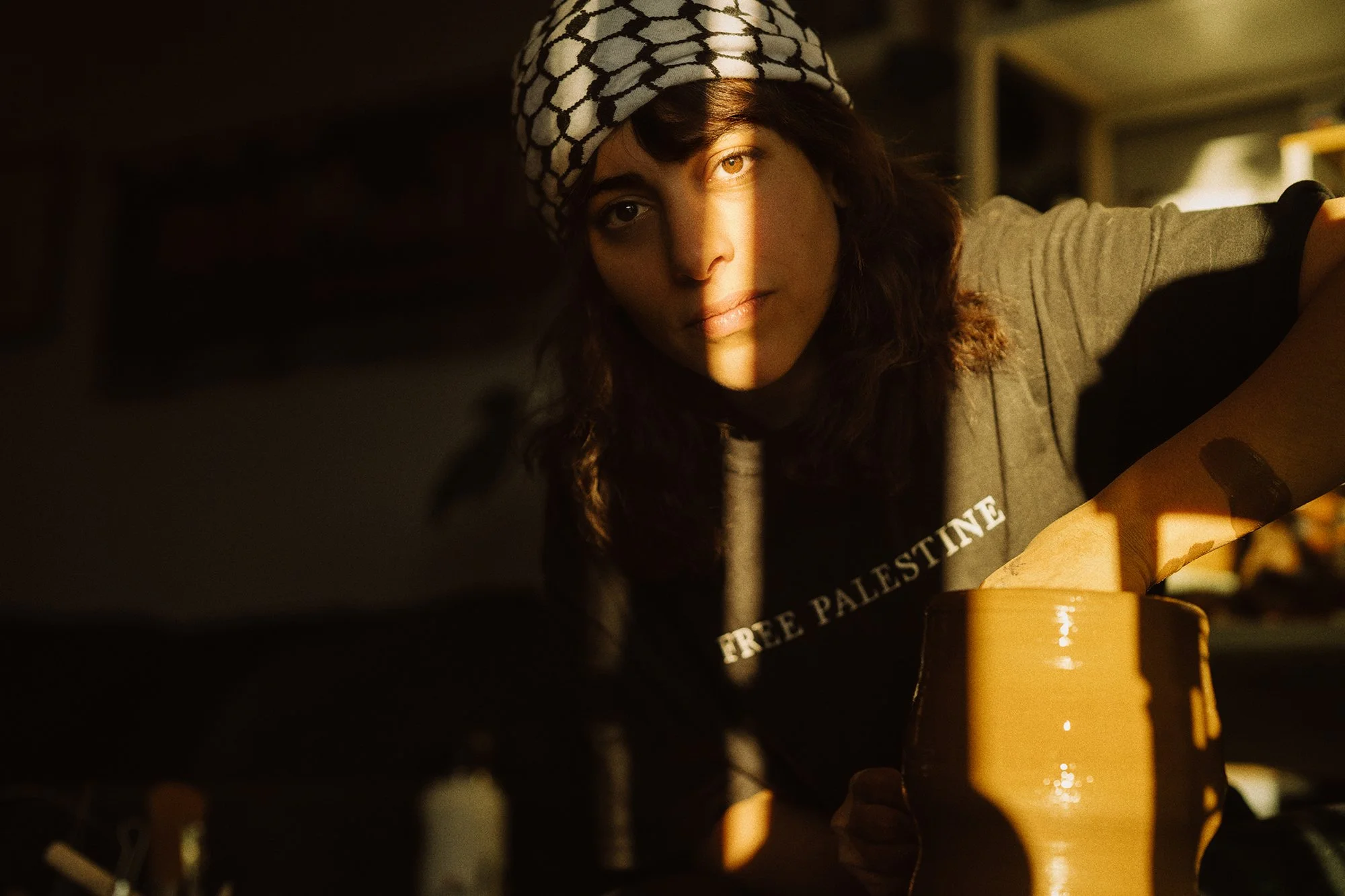 A woman with dark hair wearing a black and white headscarf and a gray t-shirt with the words "FREE PALESTINE" visible, she is looking at the camera with a soft expression, with sunlight casting a stripe of light across her face and arm.