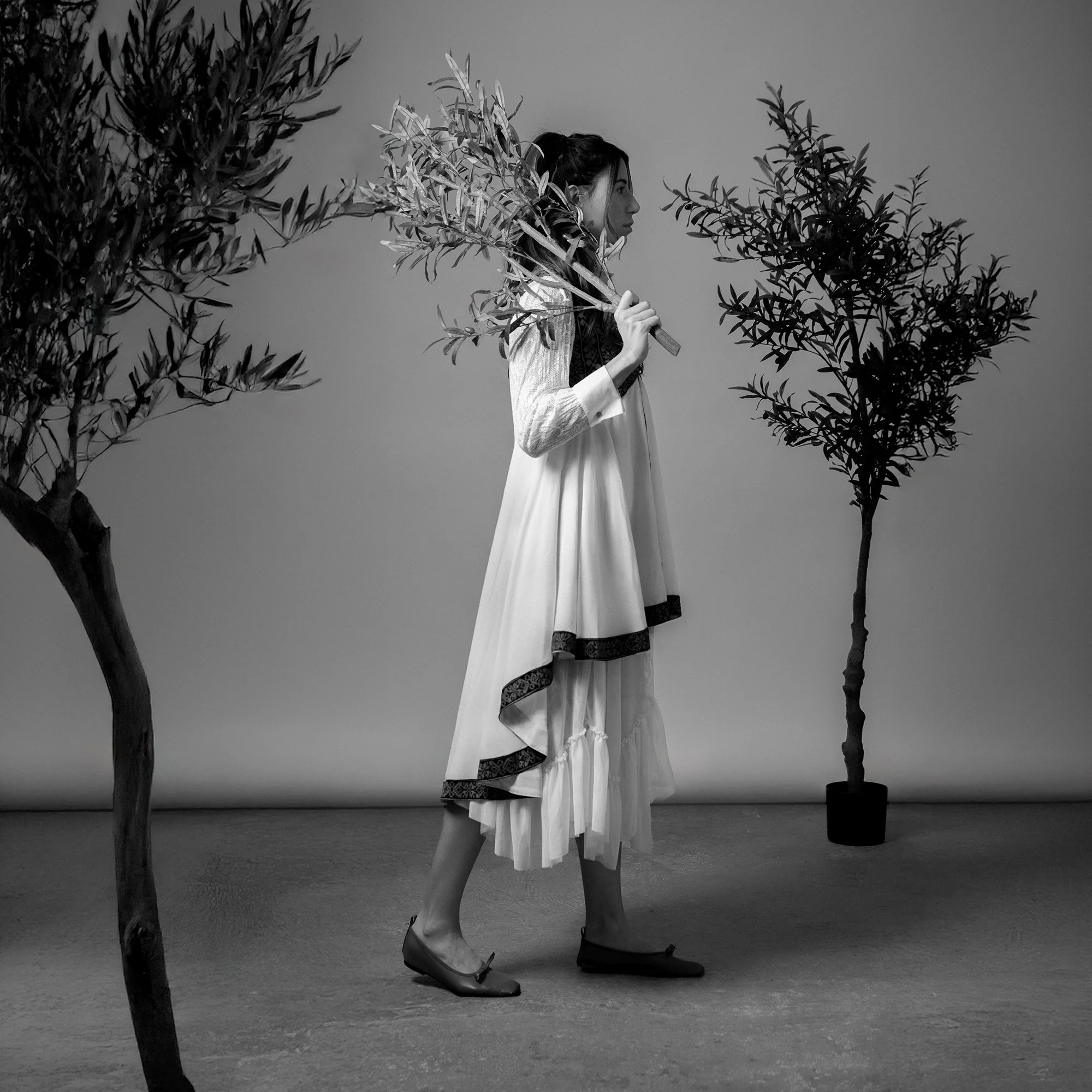 A woman dressed in traditional white and black attire holding a branch, walking between two trees in a black-and-white studio setting.