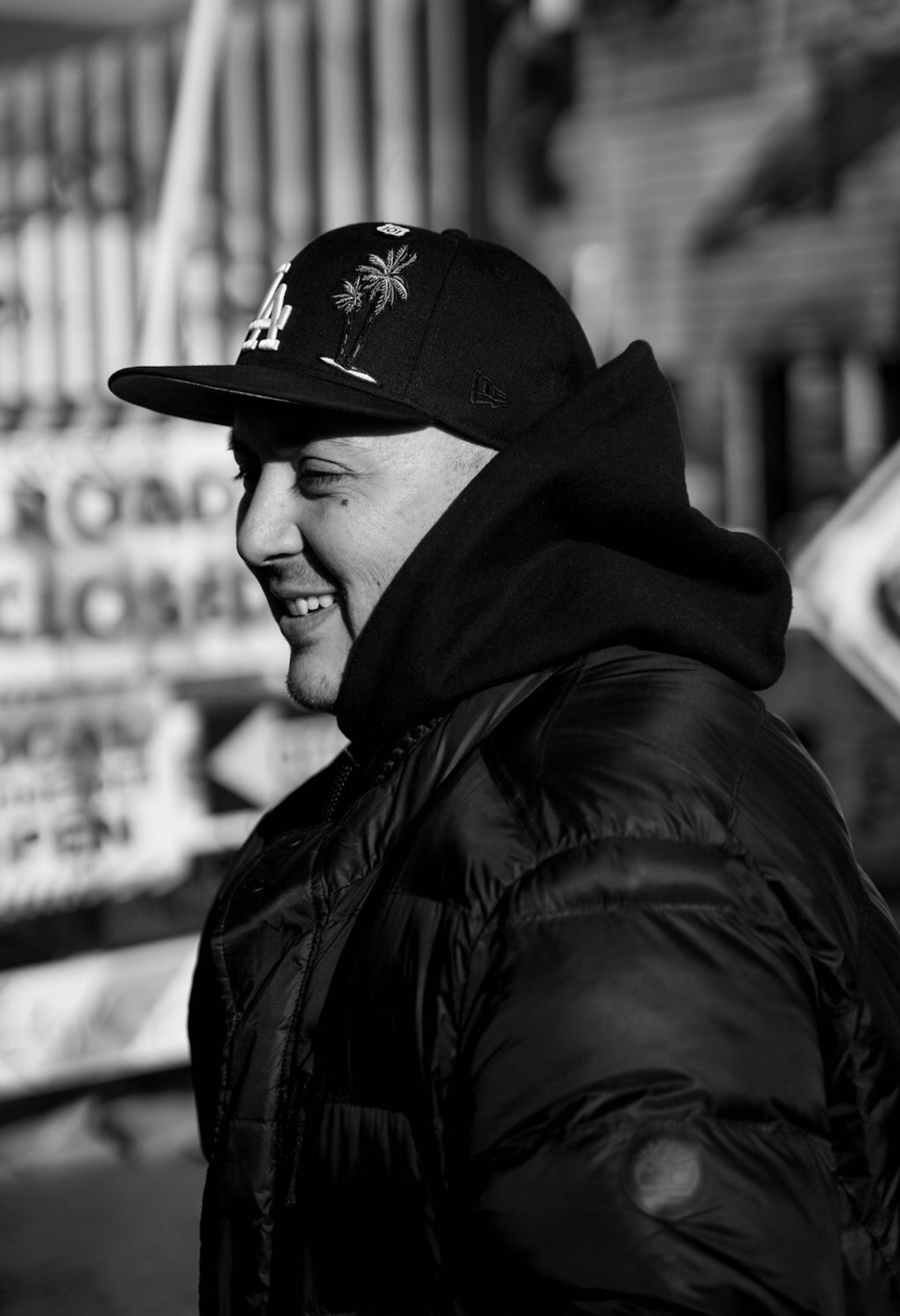 A young man smiling in profile, wearing a black baseball cap with palm trees and LA embroidered, a black hoodie, and a shiny black jacket, outdoors with blurred background.