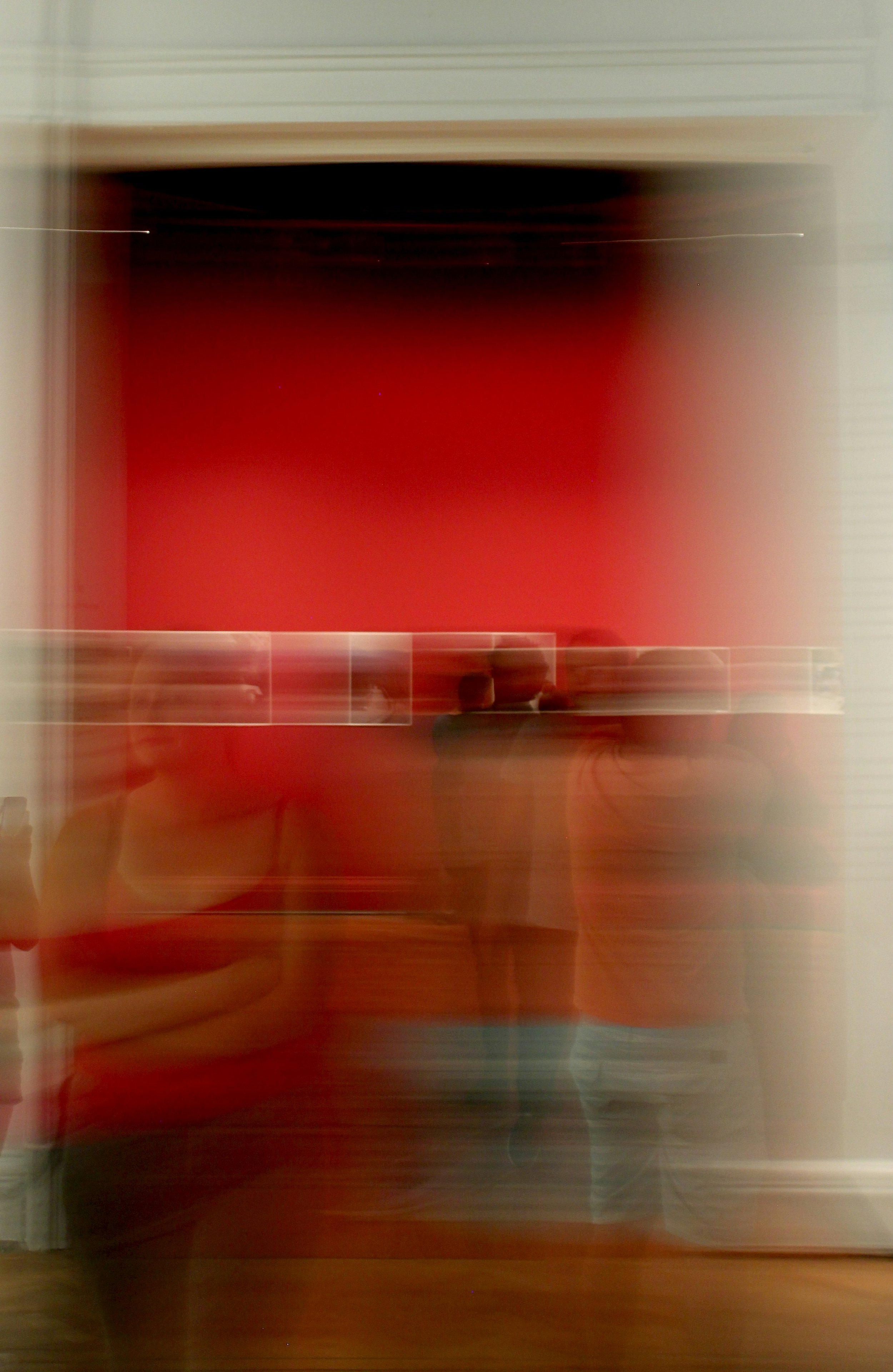 Blurred reflection of a person taking a photo in a mirror, with a red wall in the background and a horizontal tiled surface