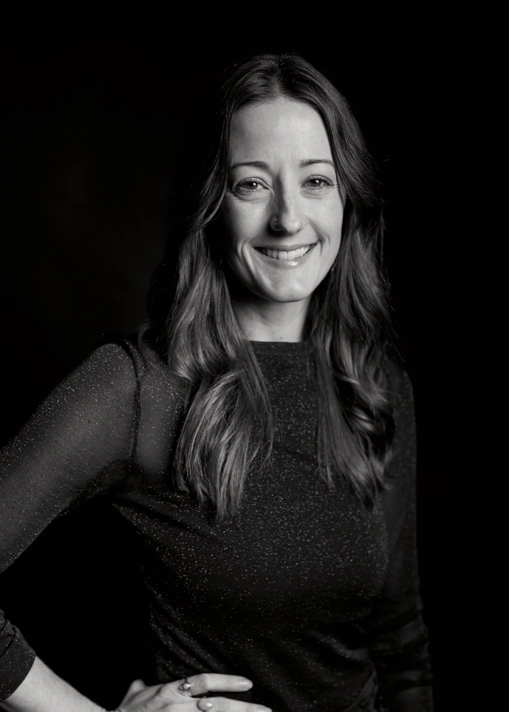 Black and white portrait of a smiling woman with wavy hair, wearing a sparkly long-sleeve top, standing against a dark background.