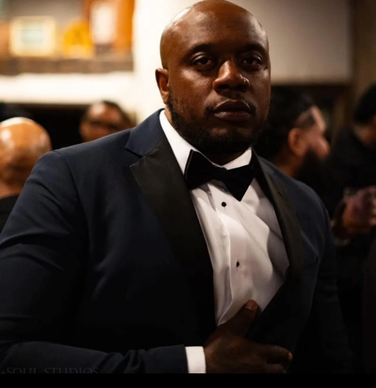 A man in a tuxedo with a black bow tie and white dress shirt, seated at a formal event, looking at the camera.